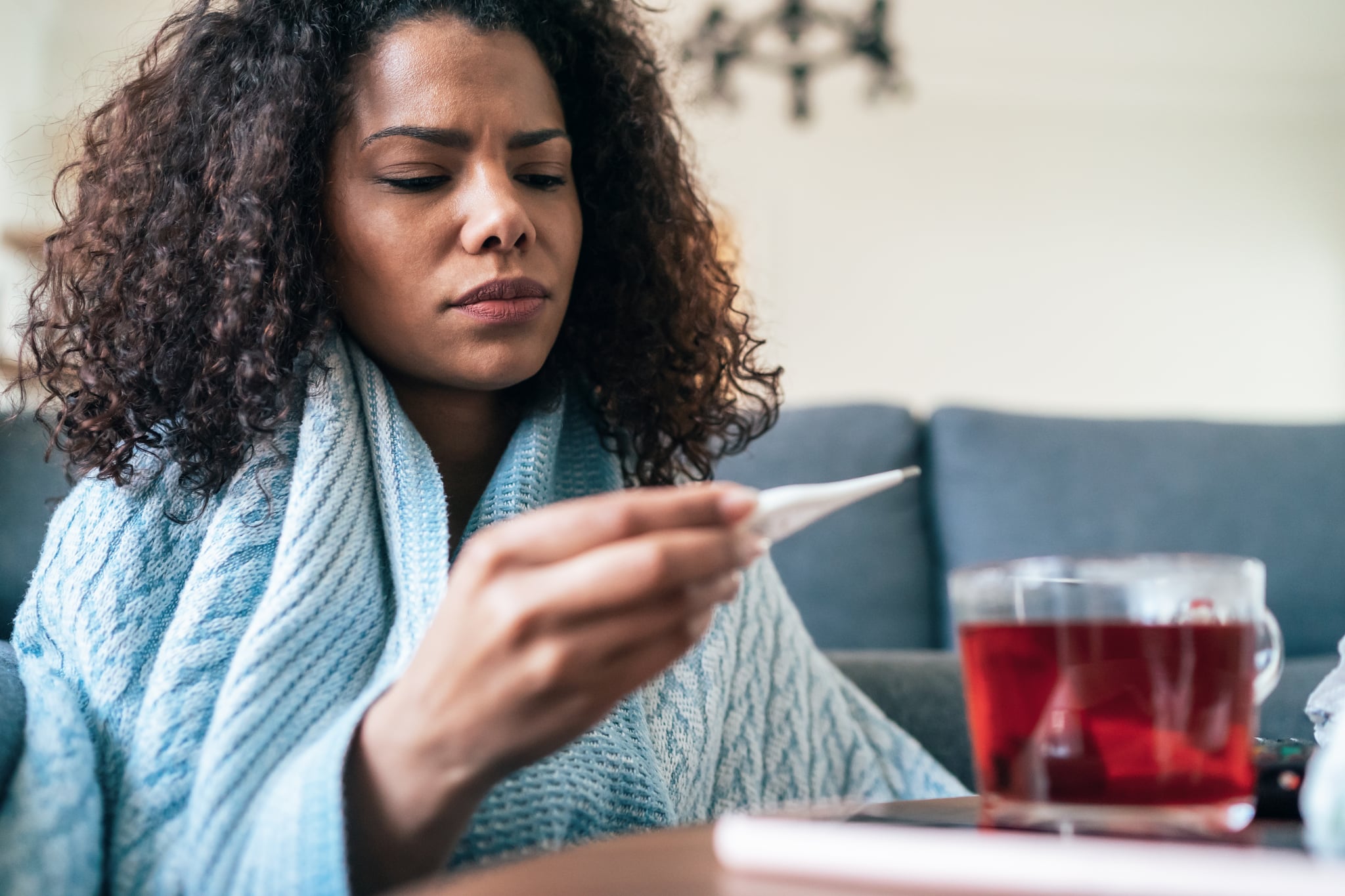 Young woman is looking at the thermometer sitting covered with blanked on the sofa at home