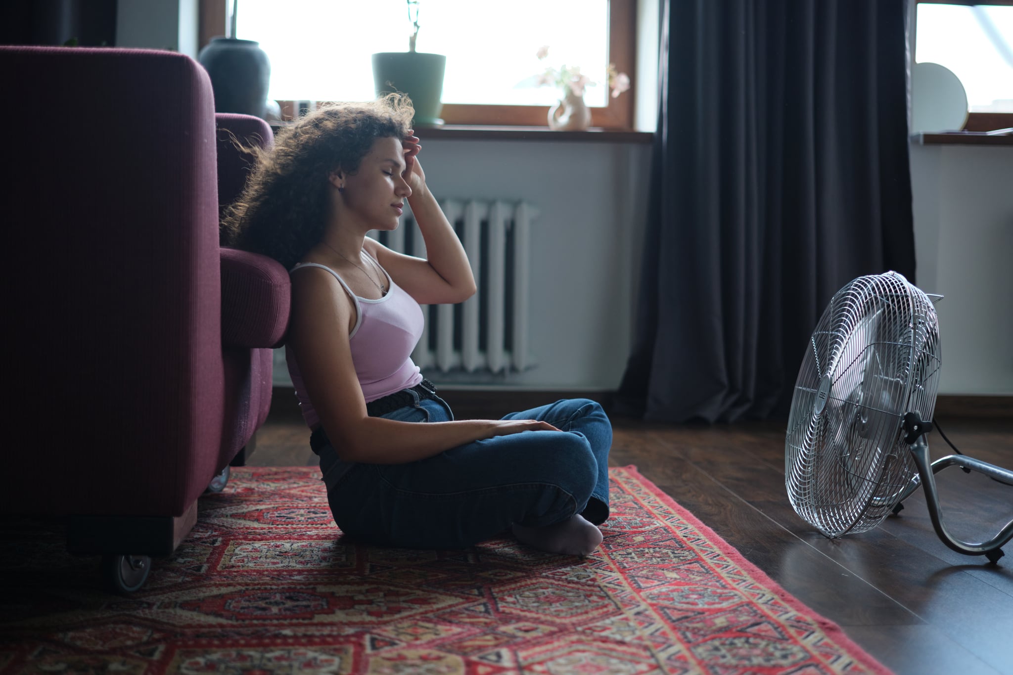 Young exhausted curly woman sitting on floor in front of electrical fan.