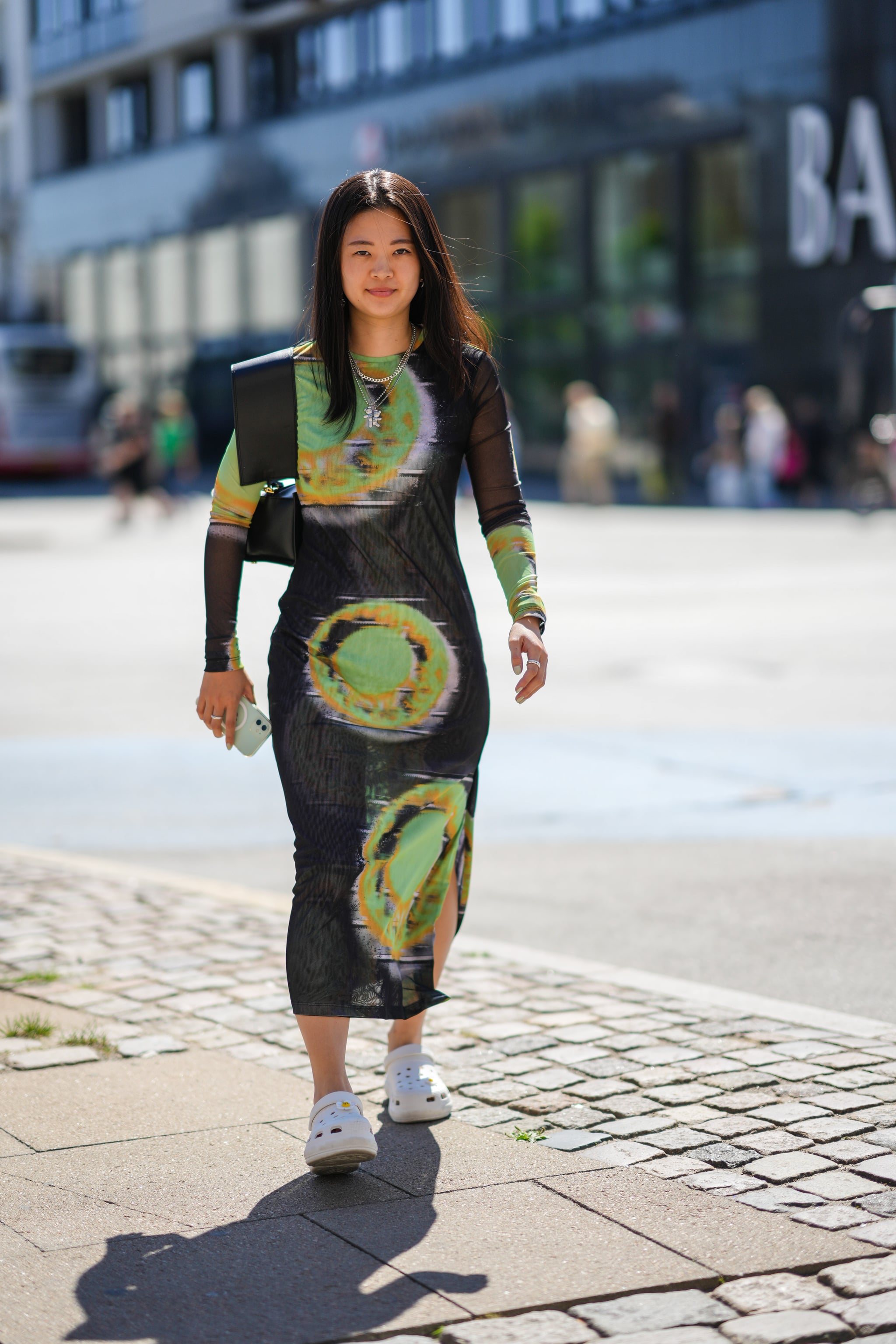 COPENHAGEN, DENMARK - AUGUST 10: A guest wears silver pendant earrings, silver chain pendant necklaces, a green / black / brown / gray tie and dye print pattern long sleeves / long dress, a black shiny leather shoulder bag from Jacquemus, white matte plastic cut-out Crocs mules, outside Holzweiler, during Copenhagen Fashion Week Spring/Summer 2023, on August 10, 2022 in Copenhagen, Denmark. (Photo by Edward Berthelot/Getty Images)