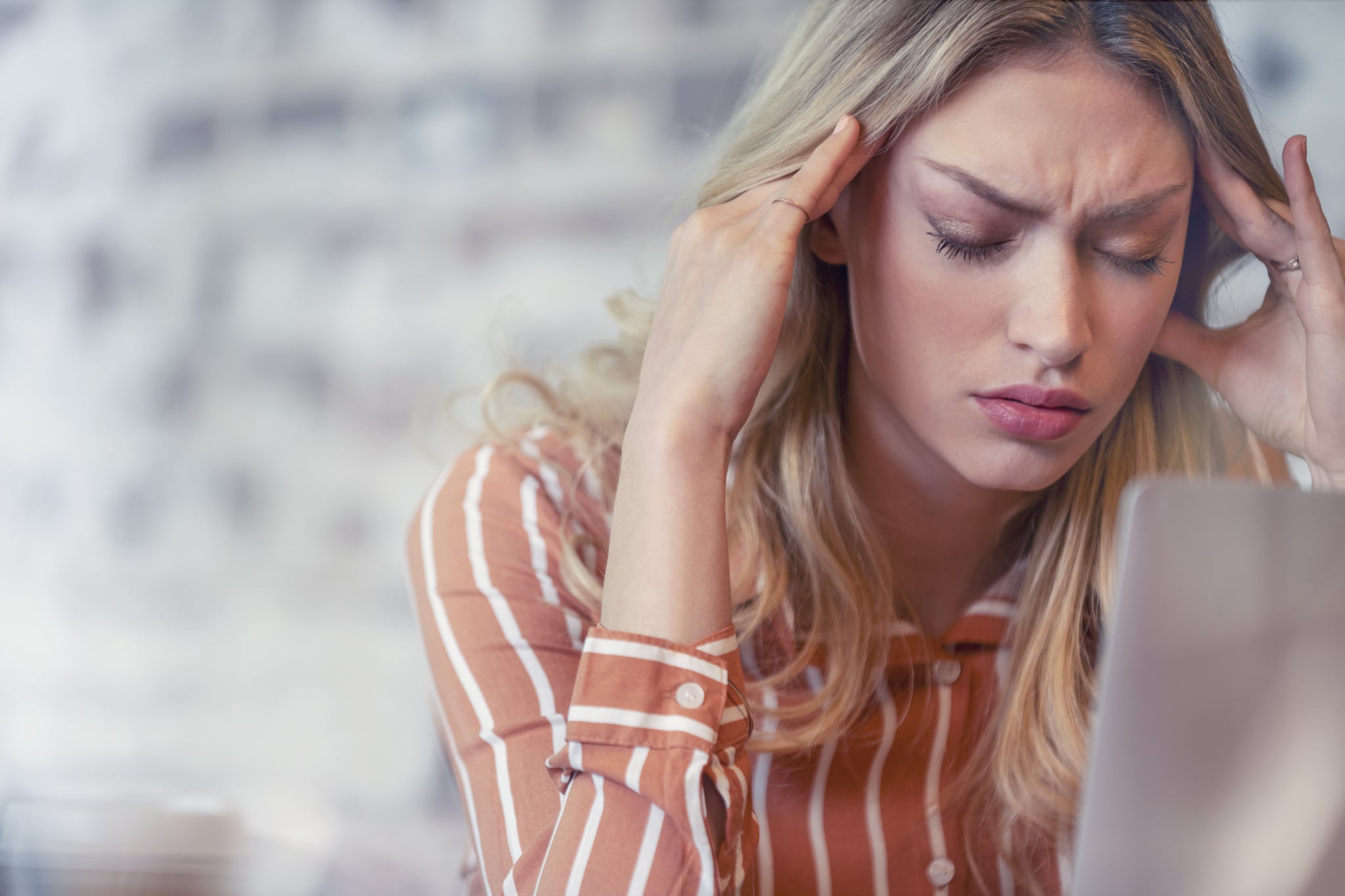 Woman stressed or worried with a headache. She is sitting at her desk using a laptop computer with her hands on her head. She has her eyes closed and is massaging her temples. Close up with copy space