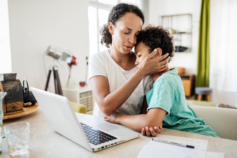 A single mom affectionately holding her young son  while she works from home in her kitchen using a laptop.