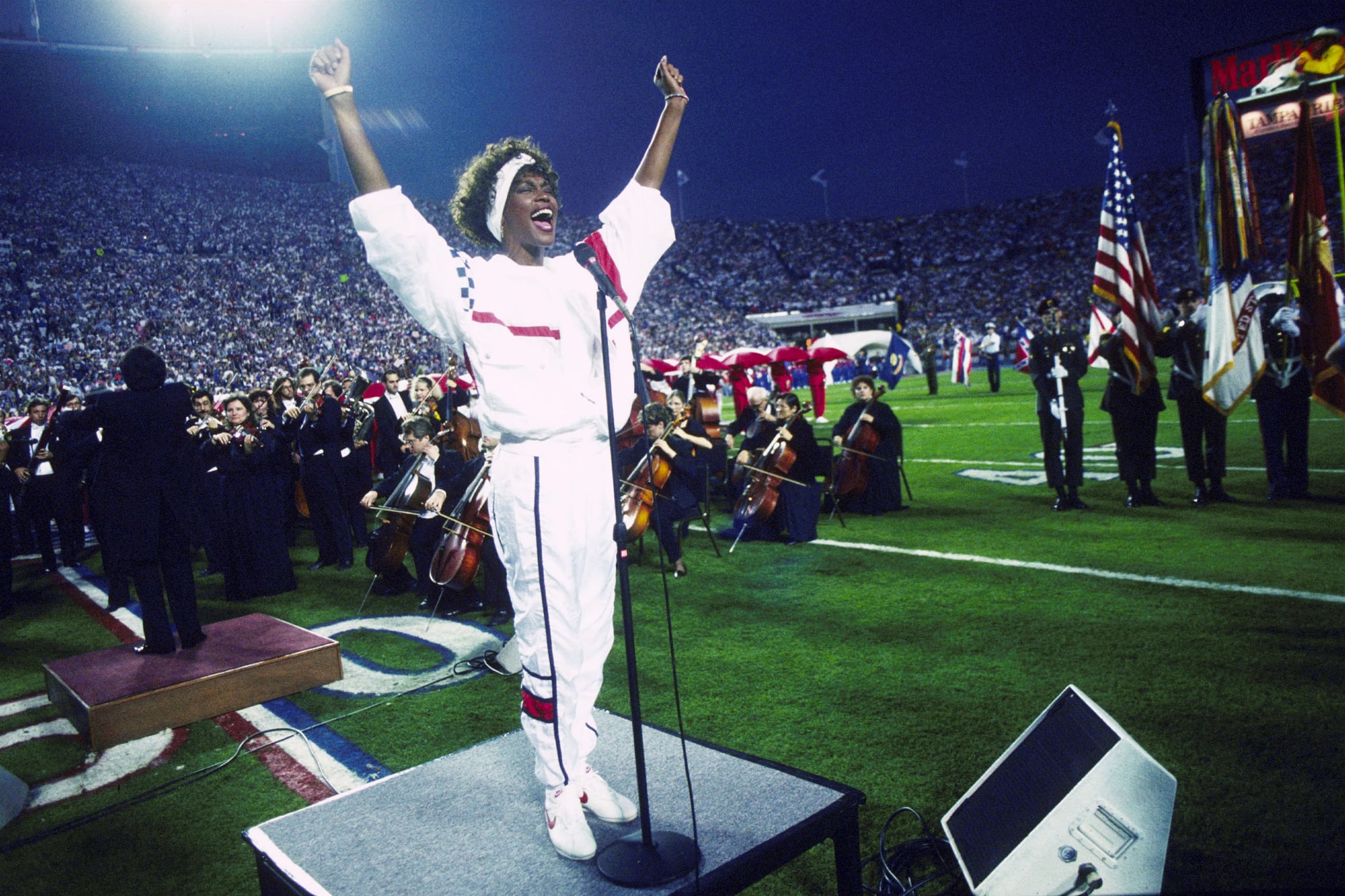 TAMPA, FL - JANUARY 27:  Whitney Houston sings the National Anthem during the pregame show at Super Bowl XXV while tens of thousands of football fans wave tiny American flags in an incredible outburst of patriotism during the Persian Gulf War on 01/27/1991.  (Photo by Michael Zagaris/Getty Images)