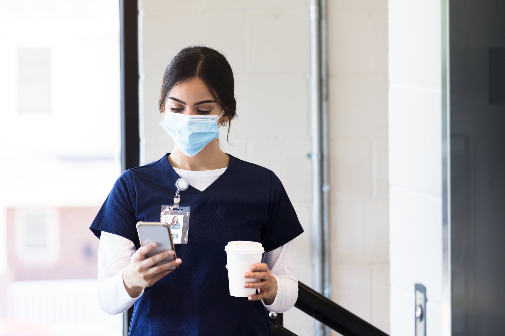 A female healthcare professional reads messages on her phone as she arrives for a hospital shift. She is wearing a protective face mask.