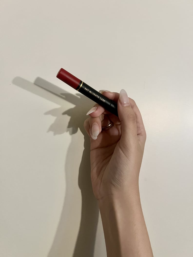 La mano de una mujer sostiene el lápiz labial Dramatique Mega Lip Pencil de Pat McGrath Labs en rojo Elson contra una pared blanca