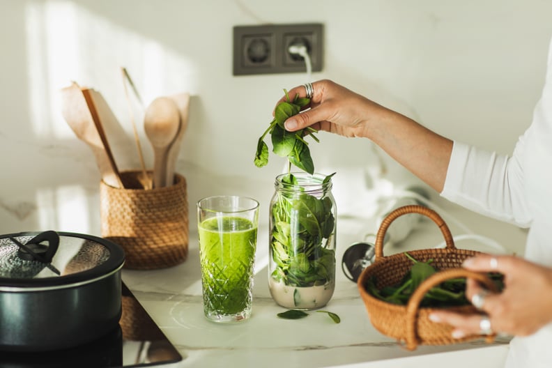 Woman making smoothie with fresh spinach and oat milk by using hand blender at home.