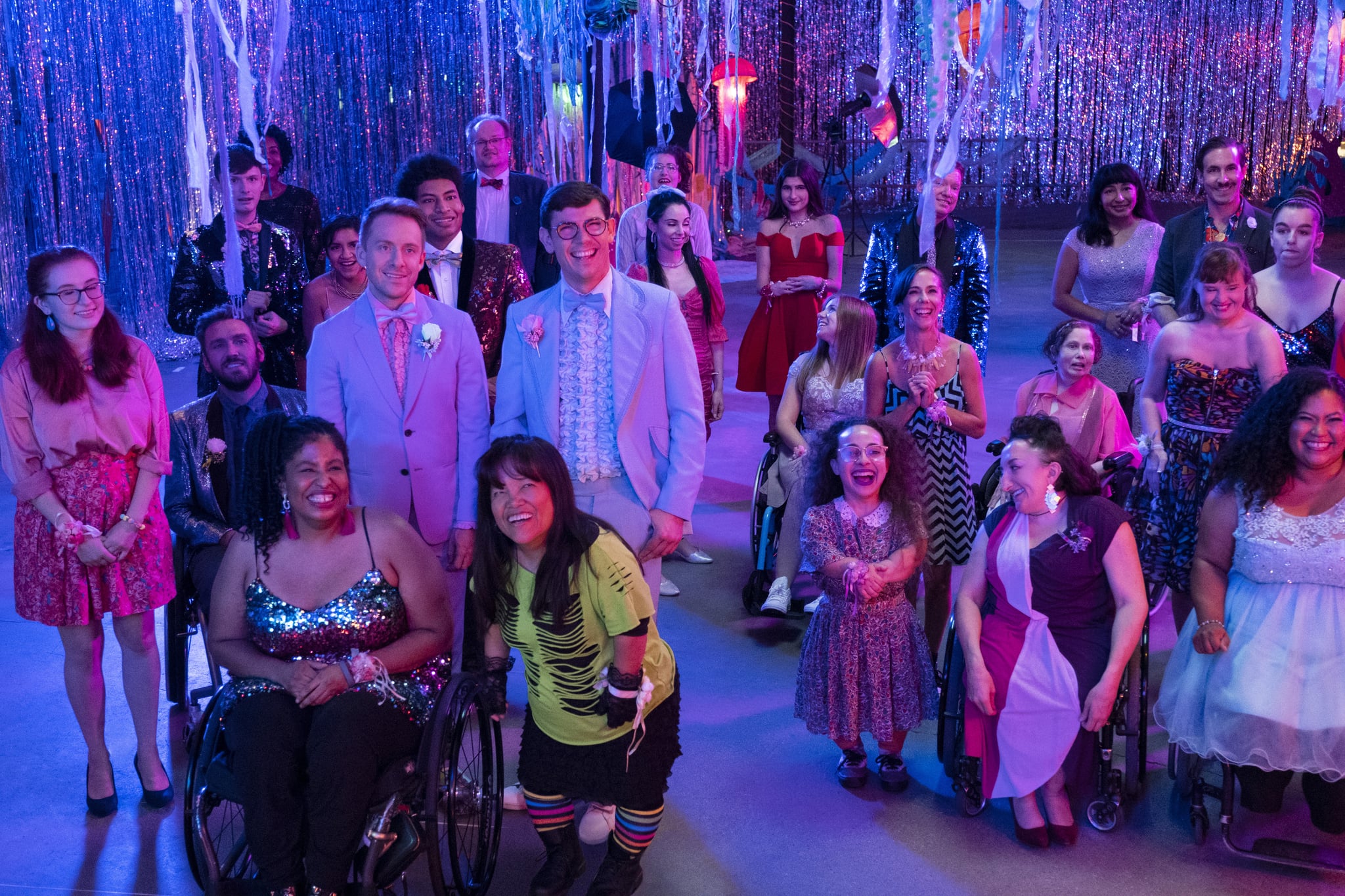 Two men in light blue tuxedos are standing in a room decorated for a party, with festive streamers hanging from the ceiling and walls. They are surrounded by men and women dressed mostly in party dresses and suits. Some of the men and women use a wheelchair, and some are people of short stature.