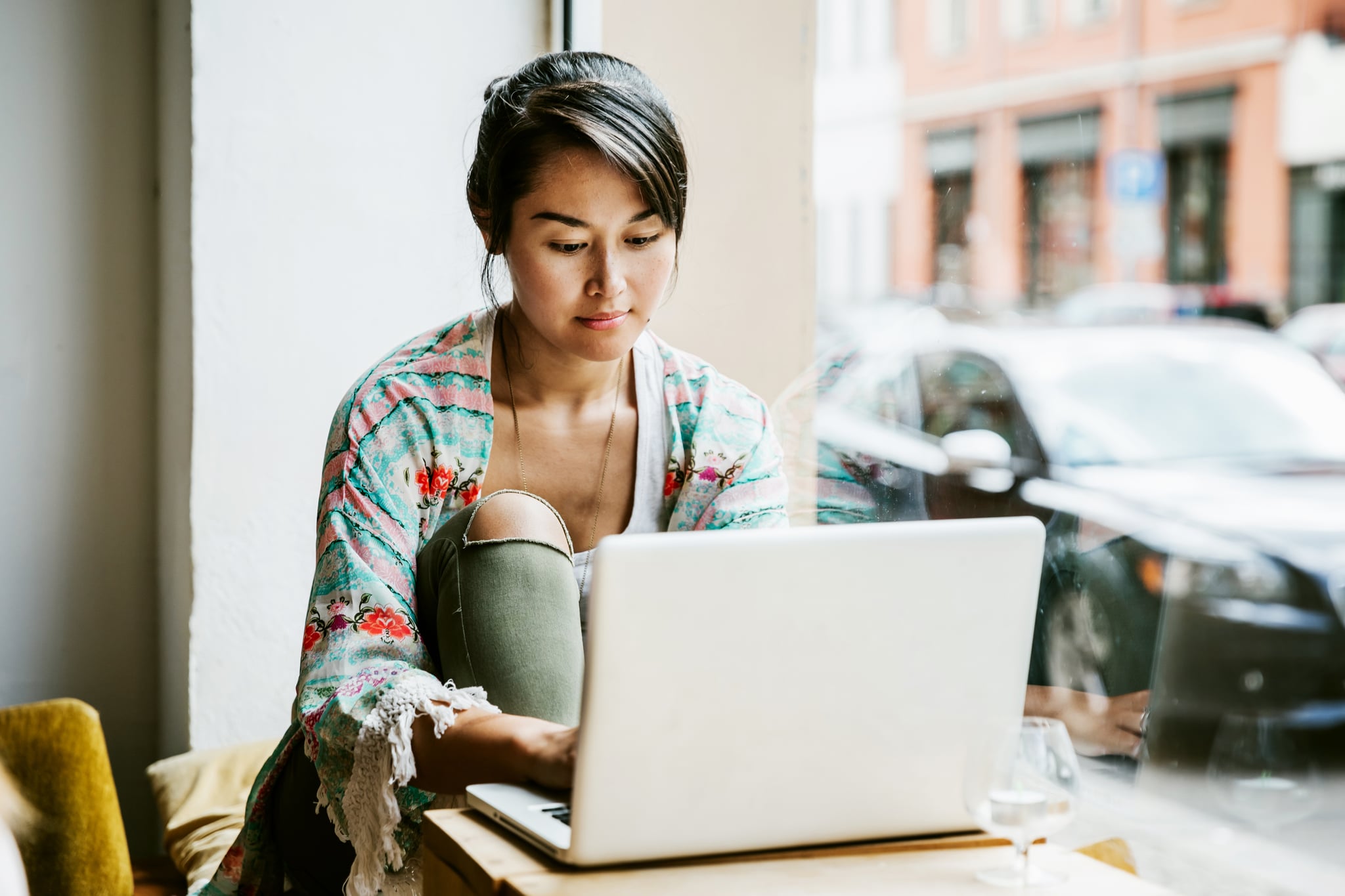 A young woman sitting at the window in a cafe is working from her laptop.