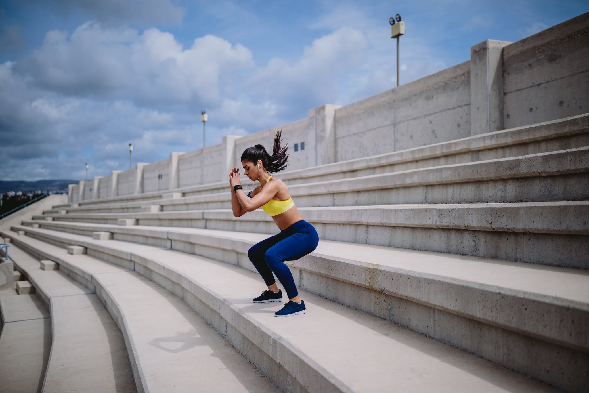 Athletic woman with smartwatch listening to music and doing jump squats on urban stadium stands