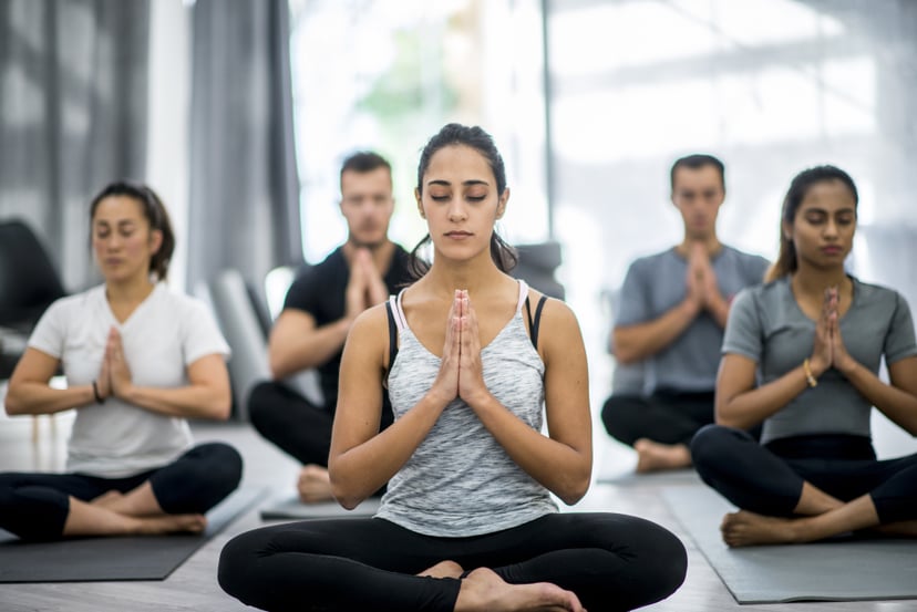 A group of adults are meditating together. They are sitting on yoga mats with their eyes closed and hands clasped together. They are in a health club.