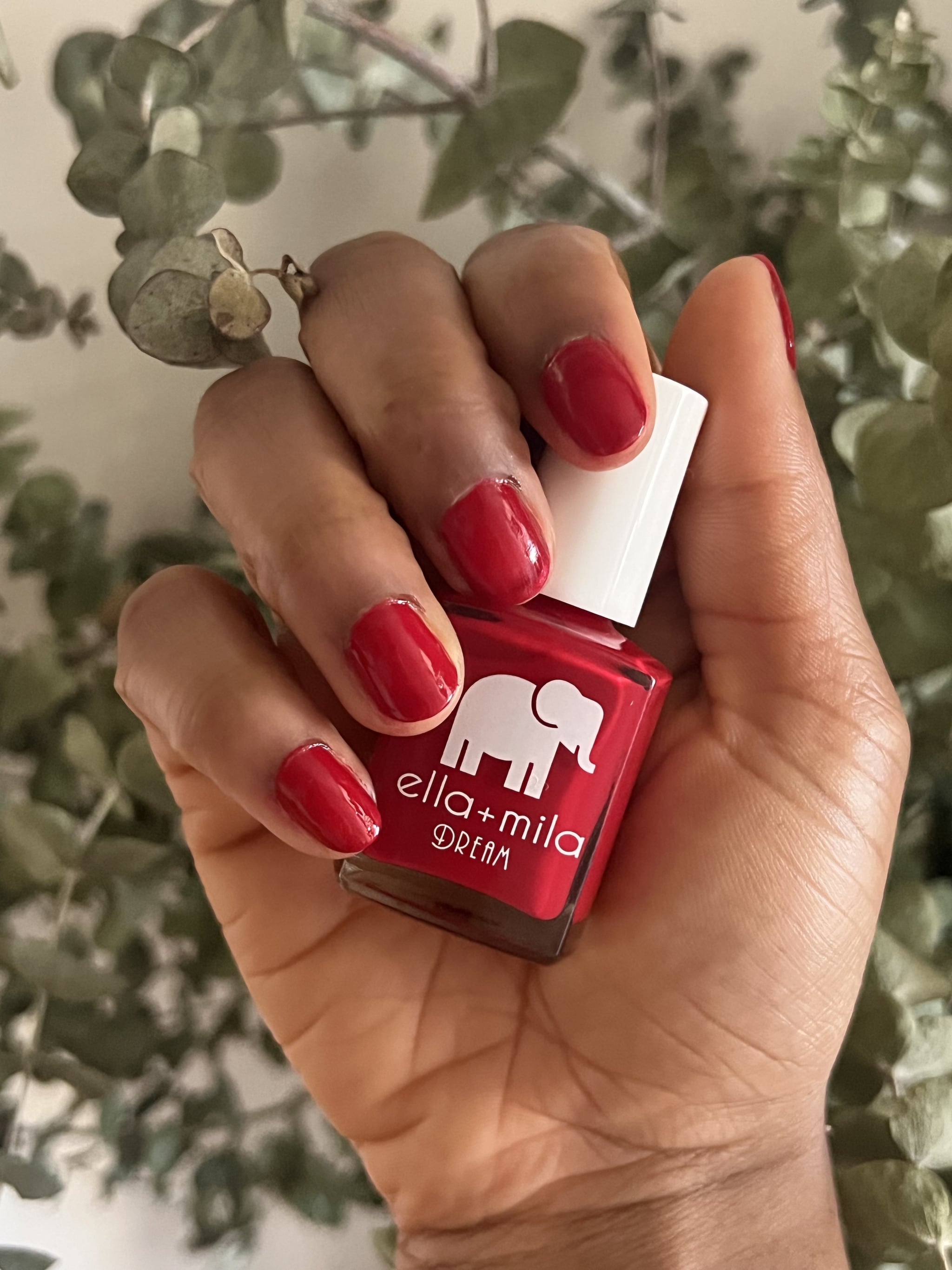 Mano de mujer sosteniendo el esmalte de uñas Ella+Mila en Unwrap Me de la colección Dream, un tono rojo intenso brillante, y luciendo el tono en sus uñas.
