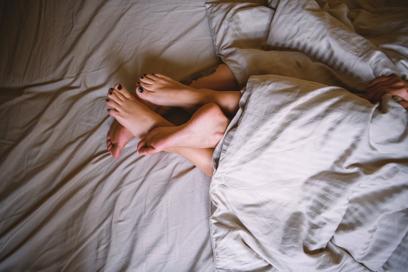 Lesbian couple lies in bed together, covered with a duvet while their legs are entwined