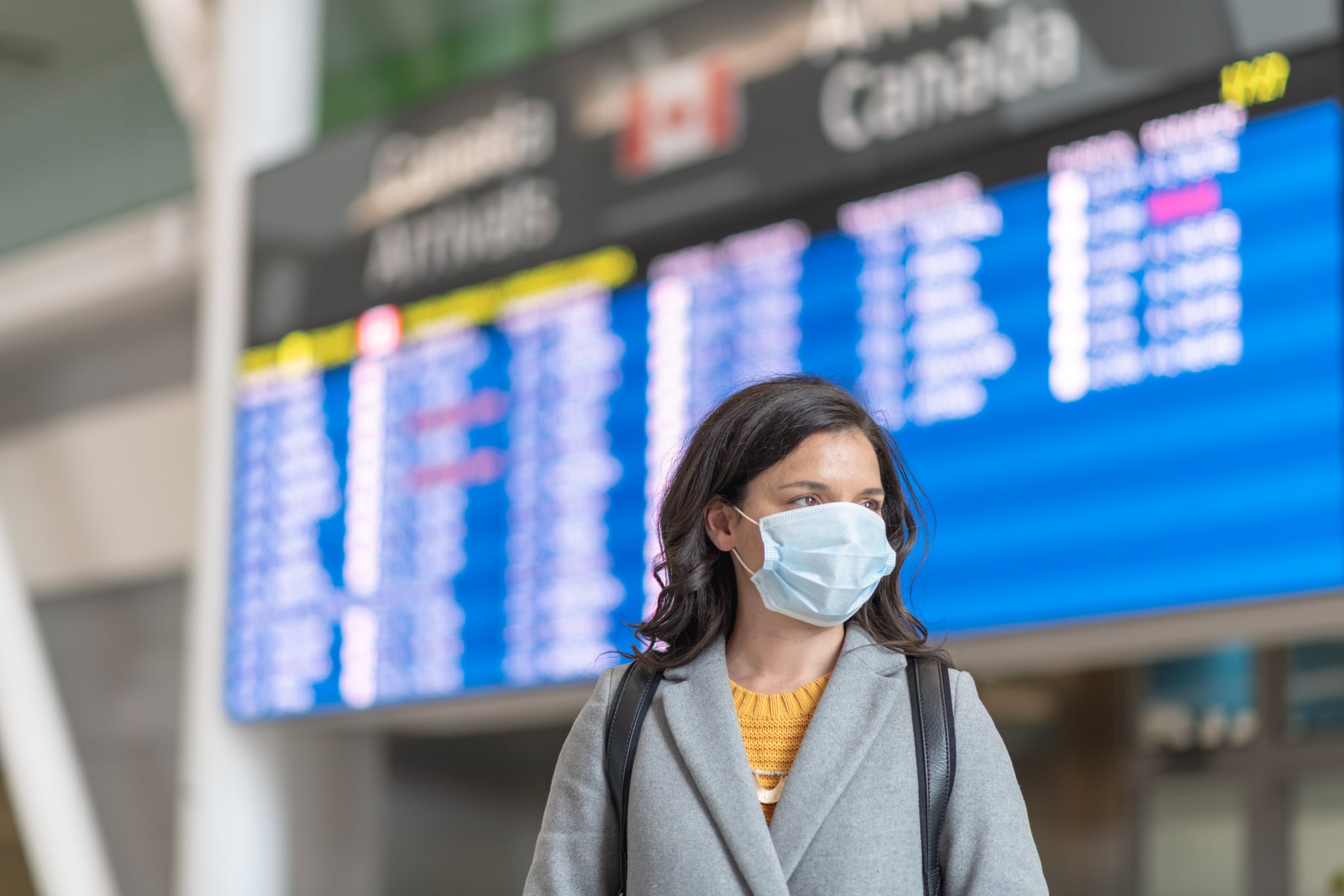 A solo travellers flight has been cancelled. She is standing in front of the departures board. She is wearing a protective face mask. Travelling during the COVID-19 pandemic.