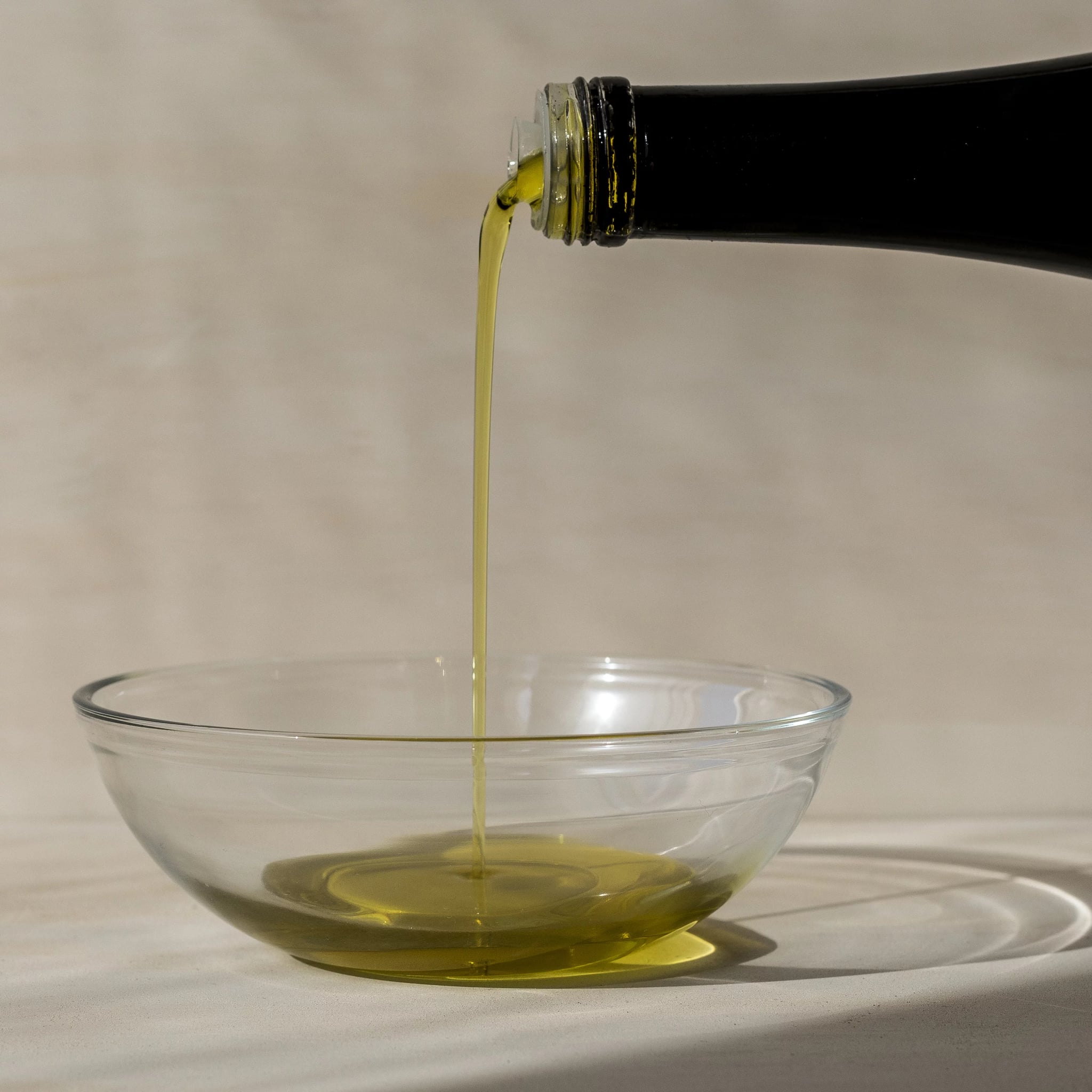 Image of the top of a black olive oil bottle with the contents being poured into a clear glass bowl. A side view photographed in natural light against a neutral background.