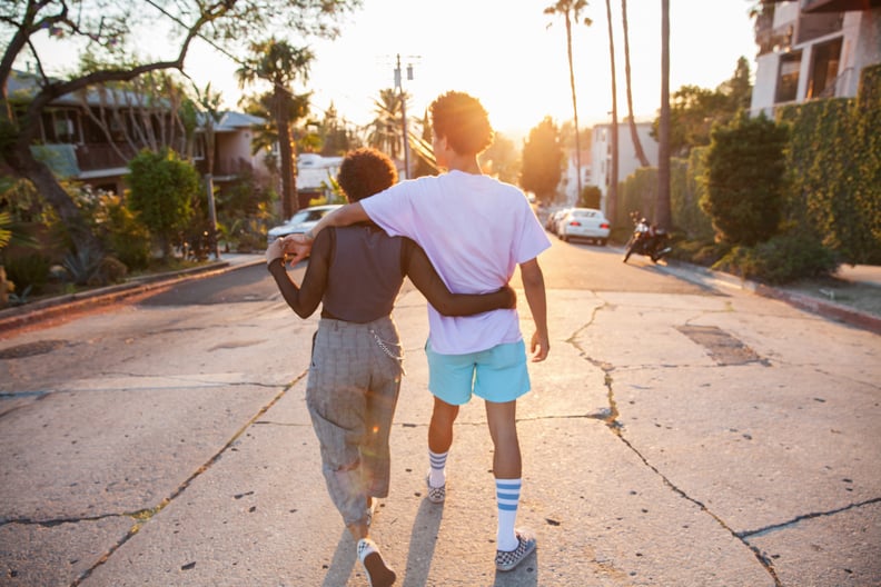 A young African American couple with their arms around each other walking down a suburban street at sunset shot from behind