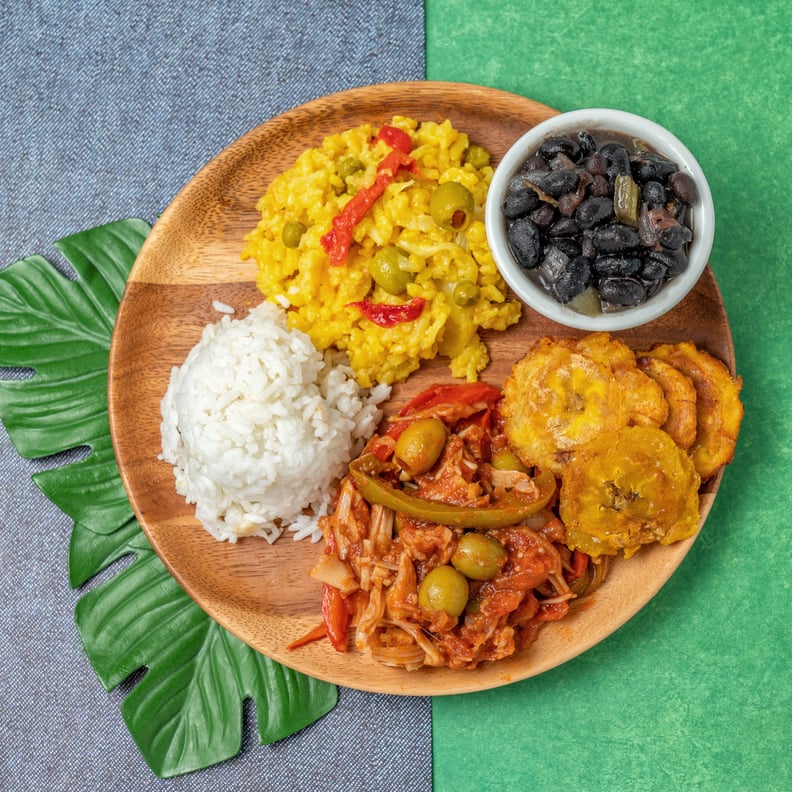 A top view of a Cuban food plate that includes yellow rice (arroz con pollo), ropa vieja, fried plantain, white rice and black beans. Shown on a wooden platter with tropical background.