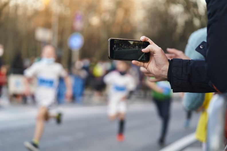 Person in black jacket holding and recording an outdoor marathon race with a phone.
