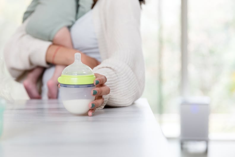 New parent picks up a baby bottle filled with infant formula while holding baby in other arm.