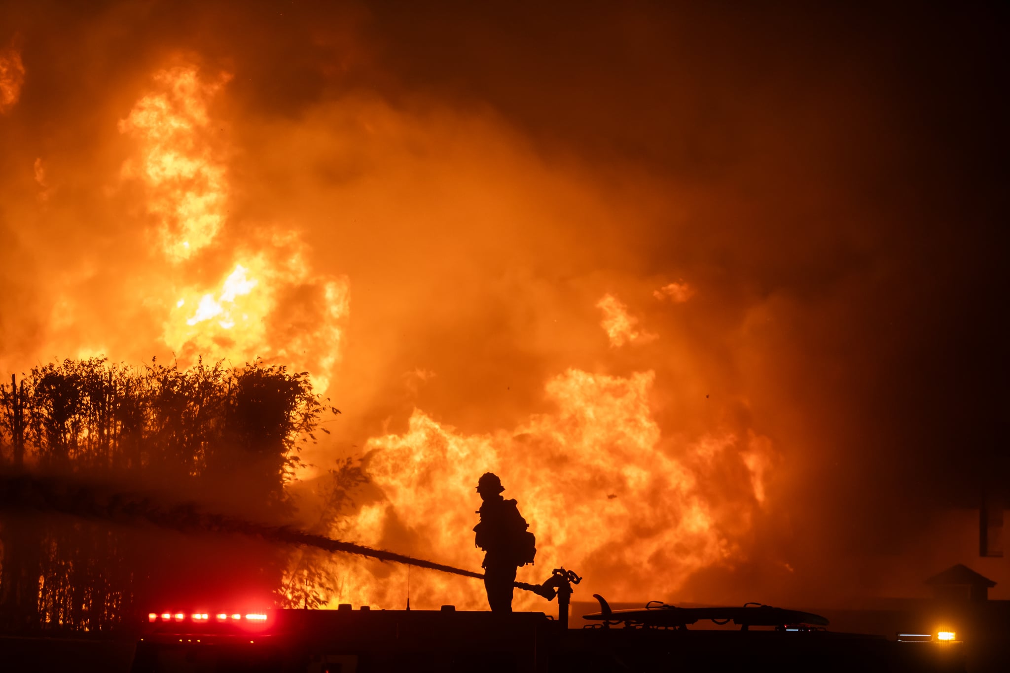 LOS ANGELES, CALIFORNIA - JANUARY 8: A firefighter stands on top of a fire truck to battle the Palisades Fire while it burns homes on the Pacific Coast Highway amid a powerful windstorm on January 8, 2025 in Los Angeles, California.  The fast-moving wildfire has grown to more than 2900-acres and is threatening homes in the coastal neighborhood amid intense Santa Ana Winds and dry conditions in Southern California. (Photo by Apu Gomes/Getty Images)