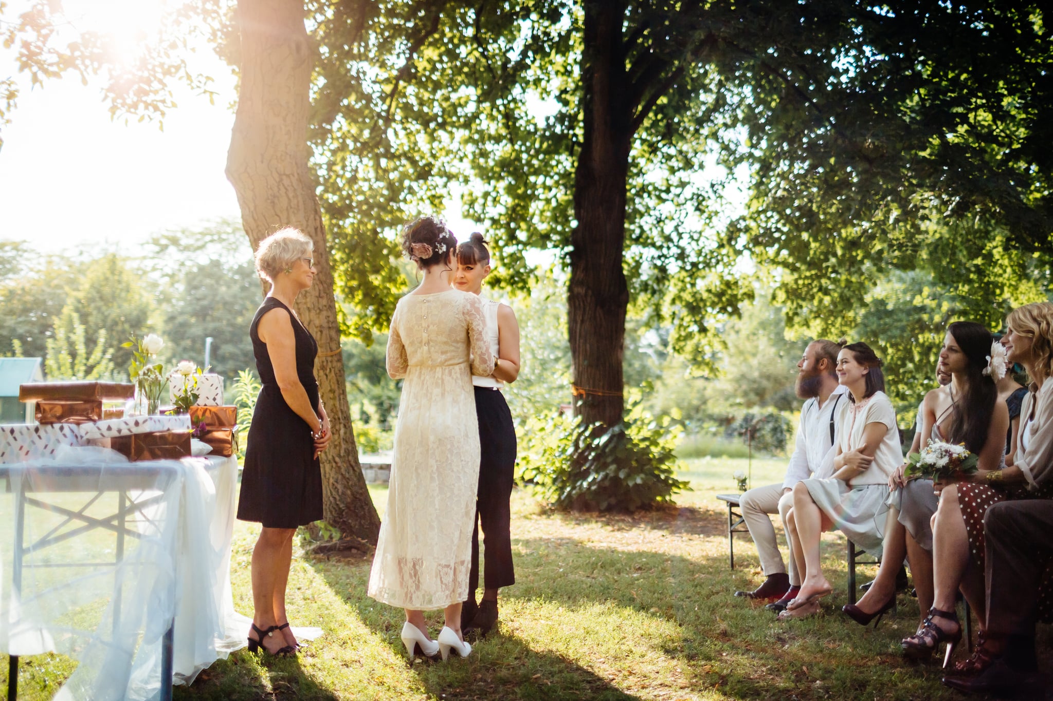 Lesbian couple holding hands in front of the altar. The wedding is outdoors and guest are seated in the foreground.