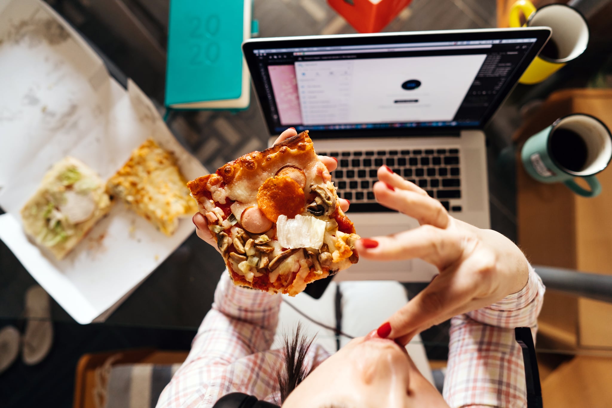 A middle-aged woman sitting in the kitchen at the glass table with laptop in headphones and holding pizza and coffee. View from above. Concepts of working at home during quarantine during a coronavirus pandemic