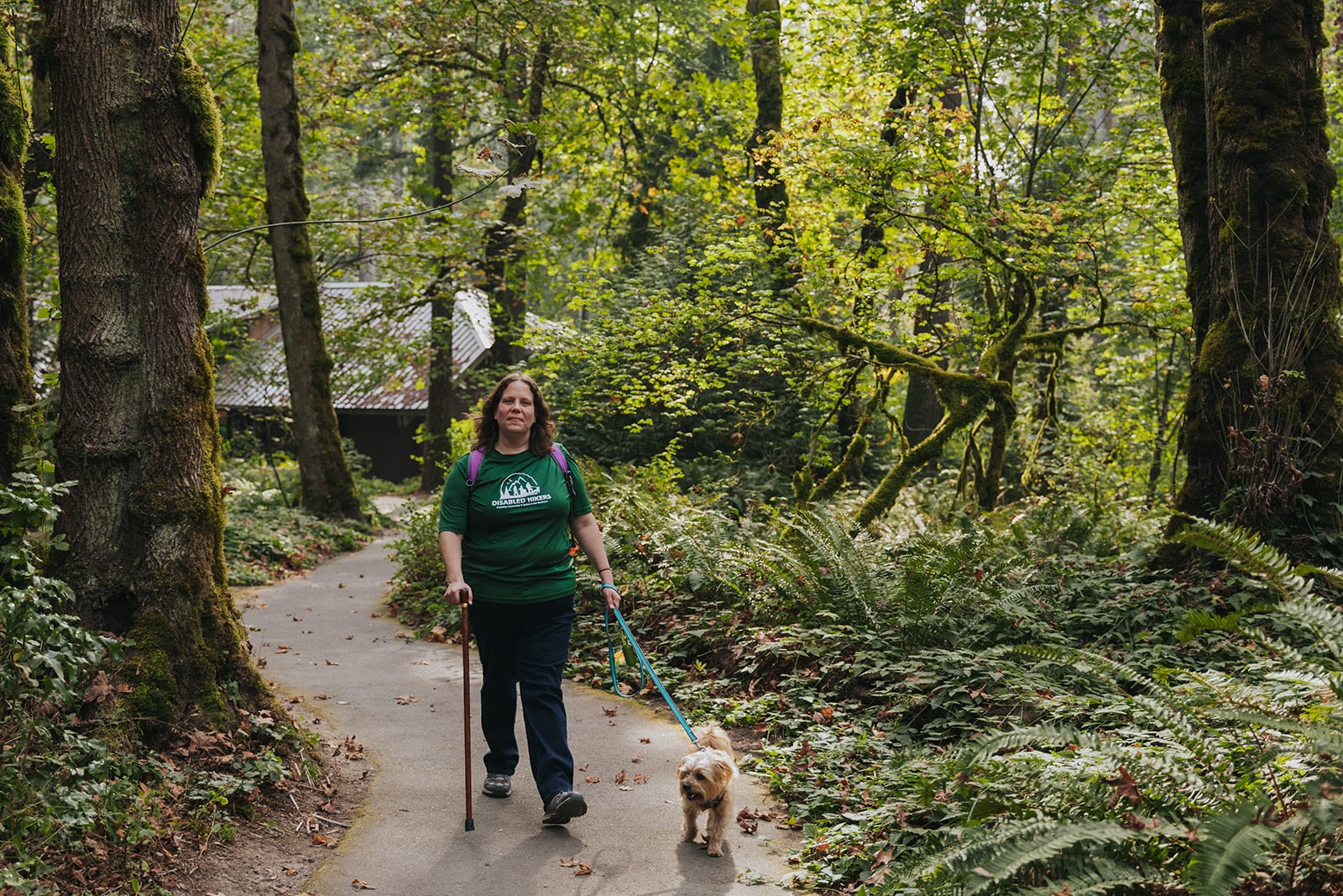 Syren Nagakyrie, a fair-skinned woman with brown hair, is wearing a green shirt and dark pants while walking her dog through a wooded trail using a walking stick.