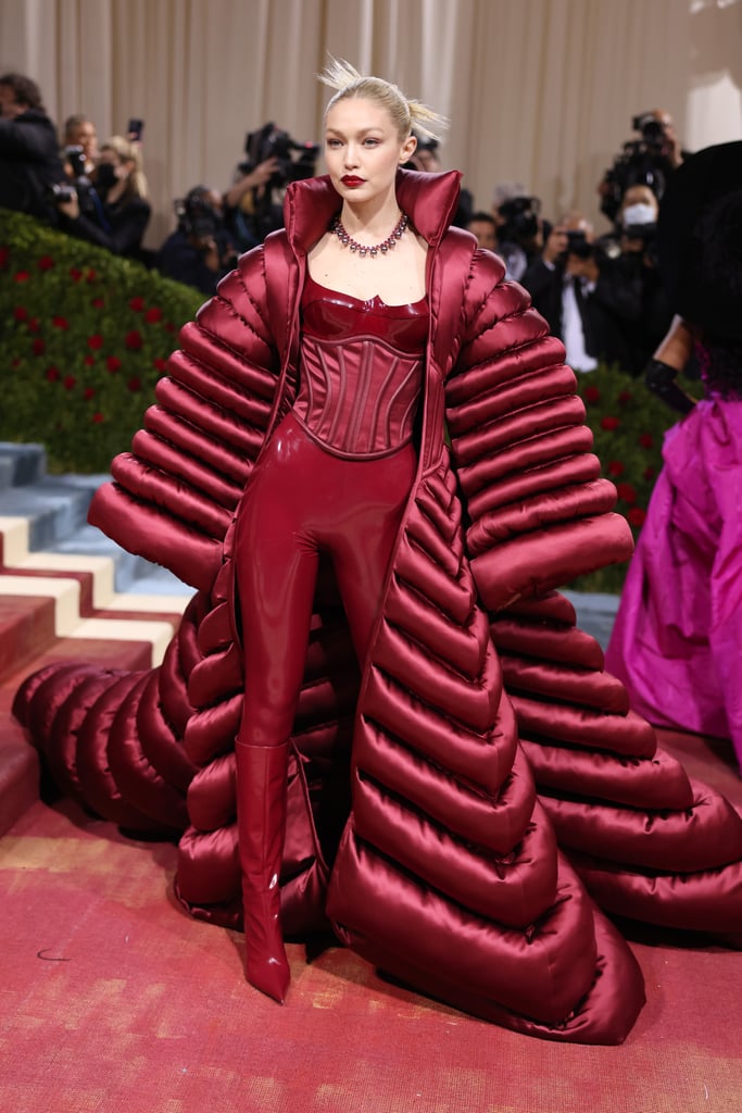 Gigi Hadid made a fiery entrance at Monday night's Met Gala. The 27-year-old supermodel donned a dramatic, floor-length parka coat over a latex look from head to toe, all in the same rich, deep shade of burgundy. Featuring a fitted corseted bodice and matching heeled boots, Hadid's vampy Versace ensemble exuded opulence on the Met Gala red carpet. Accessory wise, Hadid opted for a beaded statement necklace. And to complete her look, she styled her hair in a spiky bun and wore a complementary red lip. Leave it to Hadid to turn a winter coat into glamorous, red-carpet-ready couture. 
It's not the first time she's chosen a catsuit for the Met Gala. In 2019, the model embraced the "Camp" theme by wearing an extravagant feathery ensemble by Michael Kors, which included a matching headpiece and feather-embellished jacket. The Met Gala veteran has worn many notable looks over the years, tapping designers like Tommy Hilfiger, Versace, and Prada to craft her head-turning ensembles. At last year's event, Hadid wore a strapless gown with leather opera gloves from Prada, while in 2018, she walked the red carpet in an angelic Versace gown for the gala, where the theme was "Heavenly Bodies."  Ahead, take a closer peek at Hadid's Versace 2022 Met Gala look.

    Related:

            
            
                                    
                            

            Every Head-Turning Red Carpet Look From the 2022 Met Gala