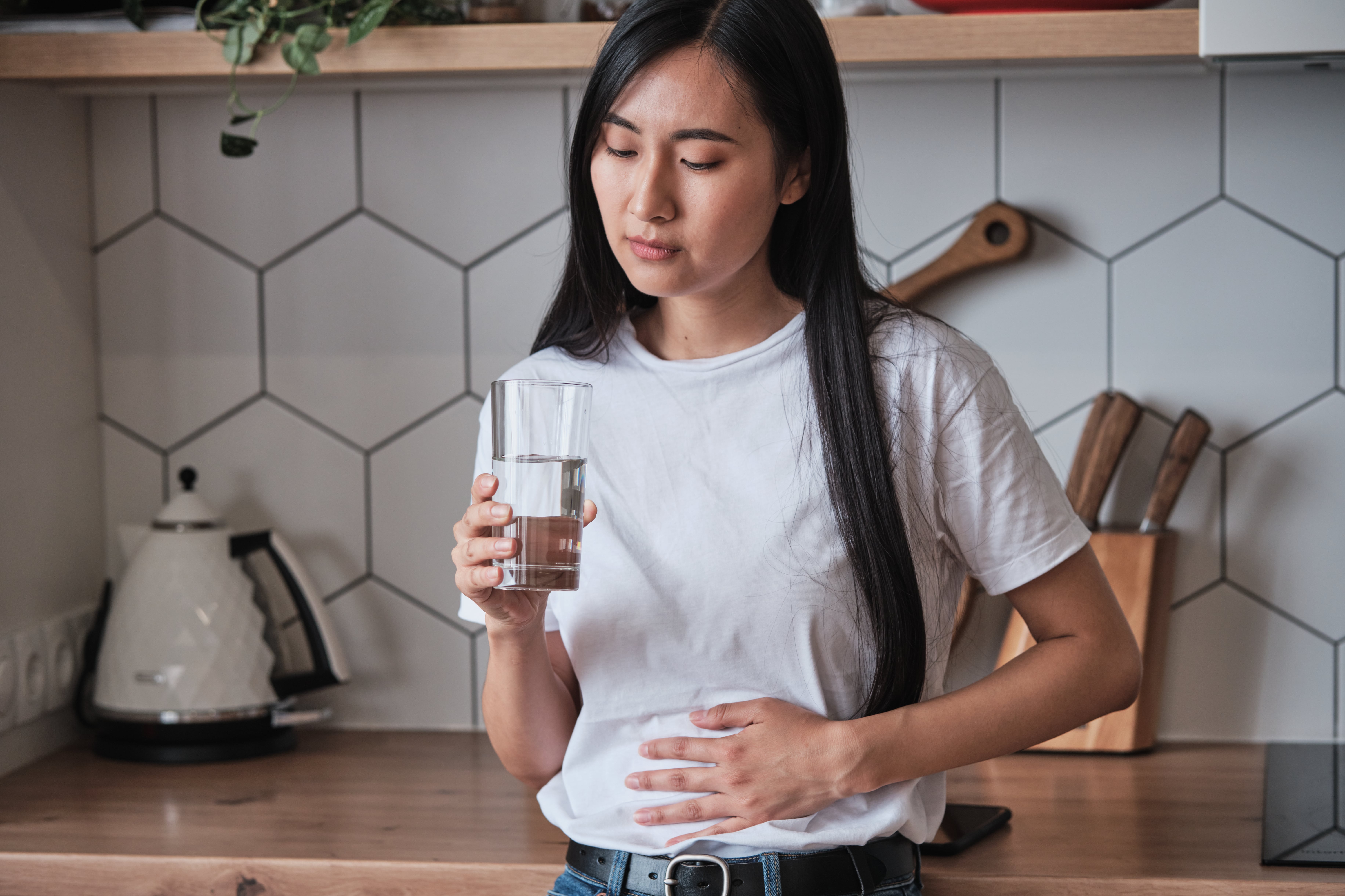 Young woman holding a glass of water and pressing her abdomen in a kitchen