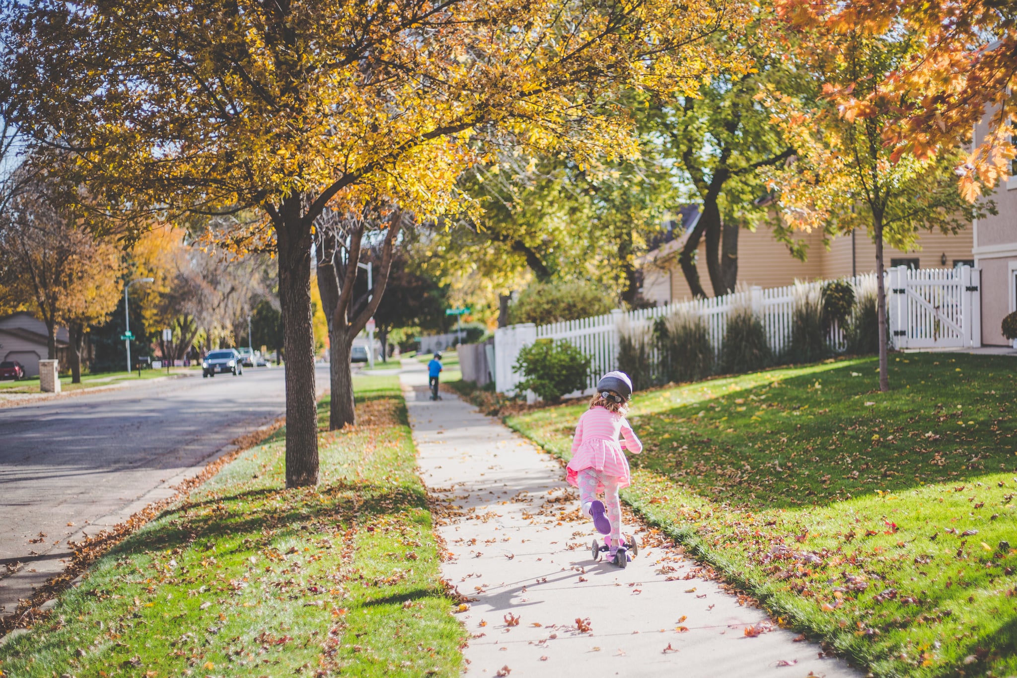 Little girl rides a scooter down a neighbourhood sidewalk on a sunny autumn day, little boy also rides far ahead of her on his own scooter.