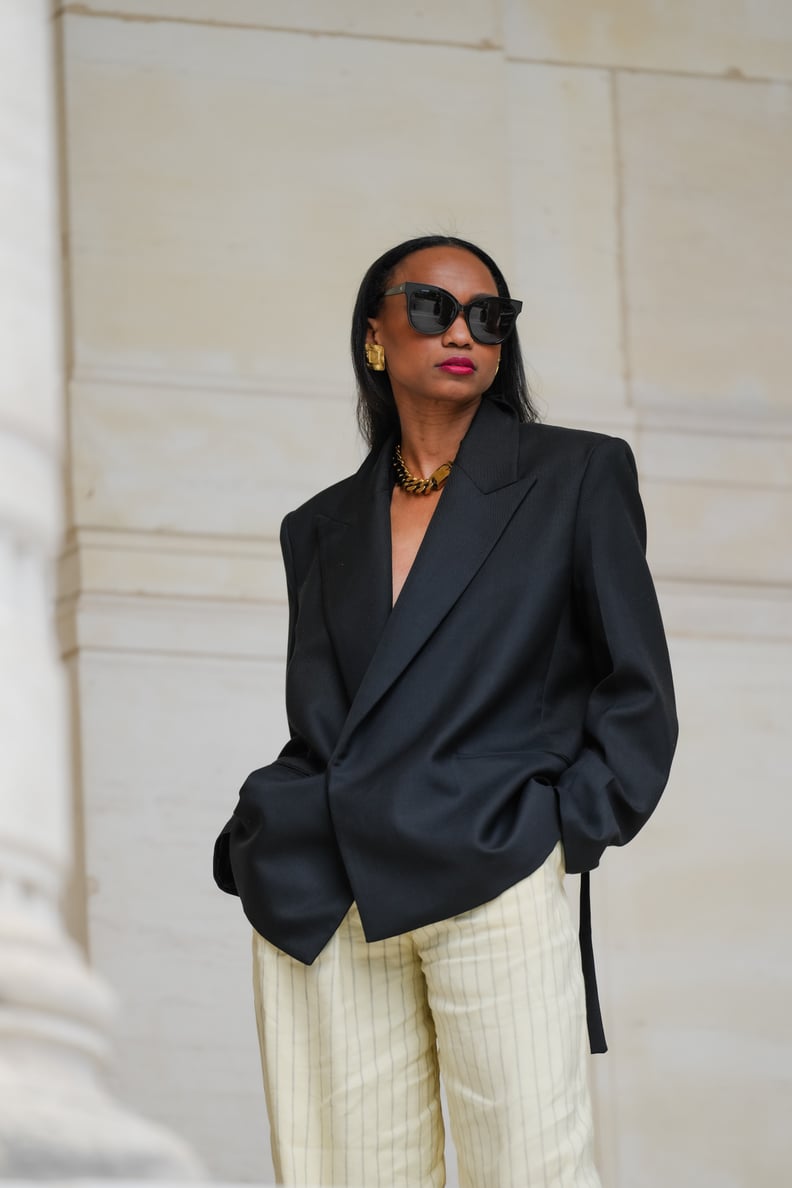 PARIS, FRANCE - MAY 12: Emilie Joseph wears sunglasses, a black oversized / structured virgin wool blend blazer jacket with lapel-collar by Victoria Beckham for Mango, pinstripes / striped linen high-rise trousers / dad flared wide-leg pants in butter pal