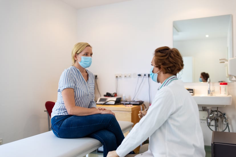 Woman wearing face mask sitting on examination table talking with female doctor holding an injection. Doctor discussing with patient before giving a vaccine to her.