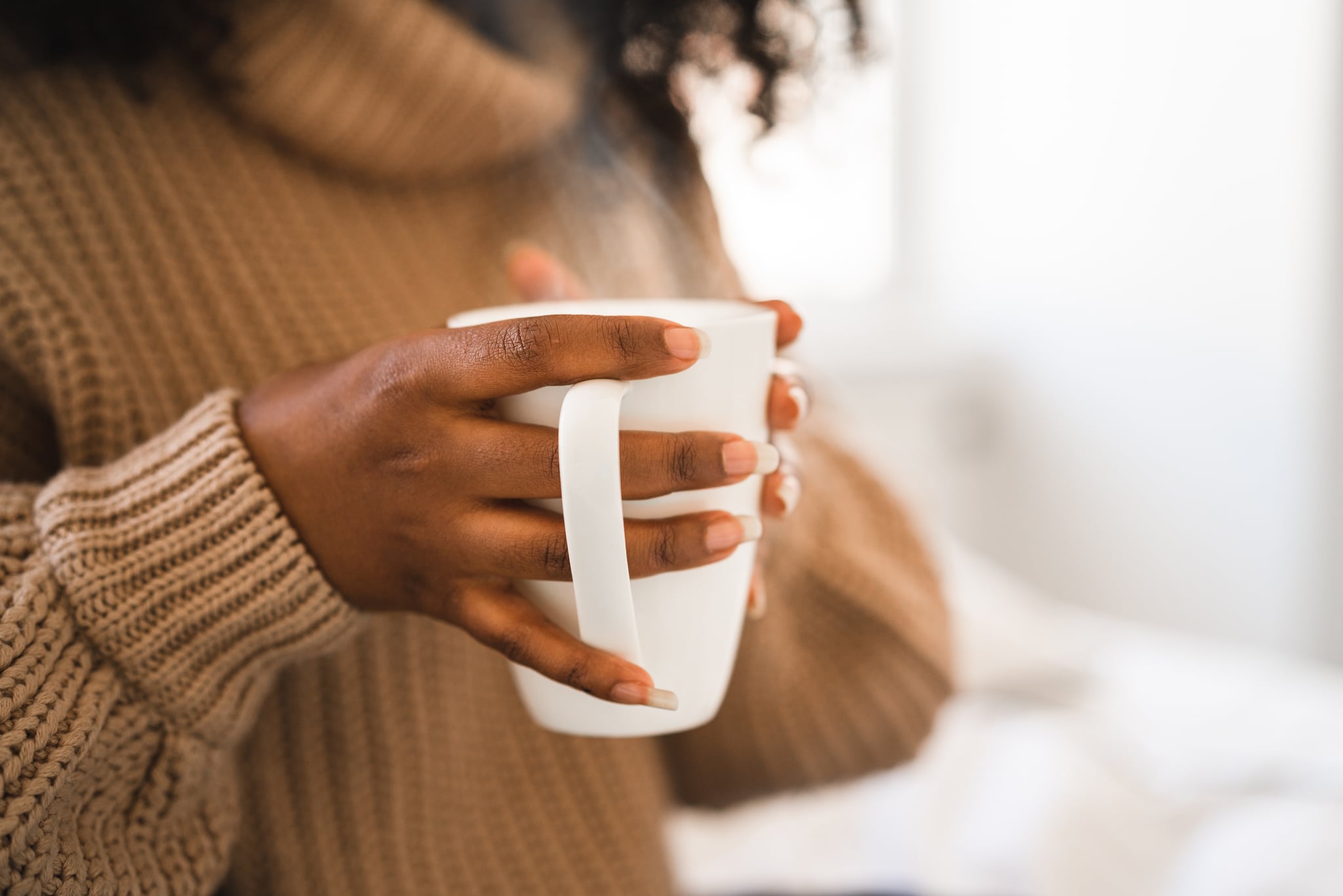 A female university student is sitting in her bed, studying on her laptop. She is working from home for social distancing purposes. She is holding a cup of hot coffee and watching a web conference.