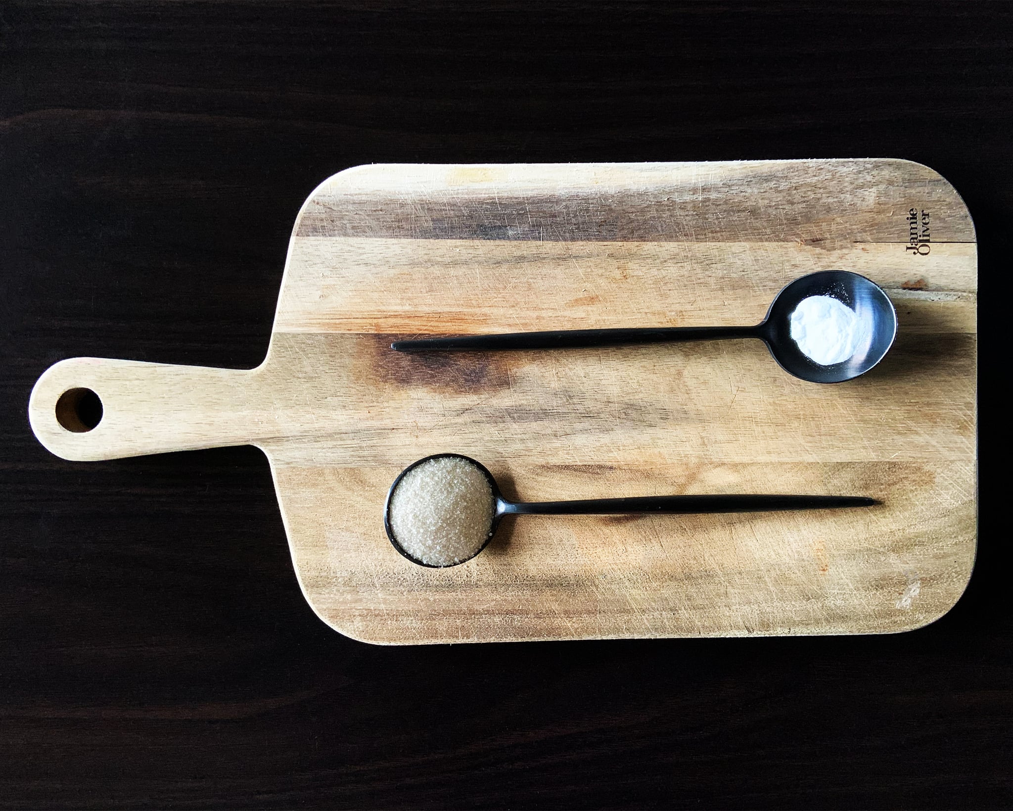 Two black spoons on a cutting board, one has sugar the other bicarbonate of soda