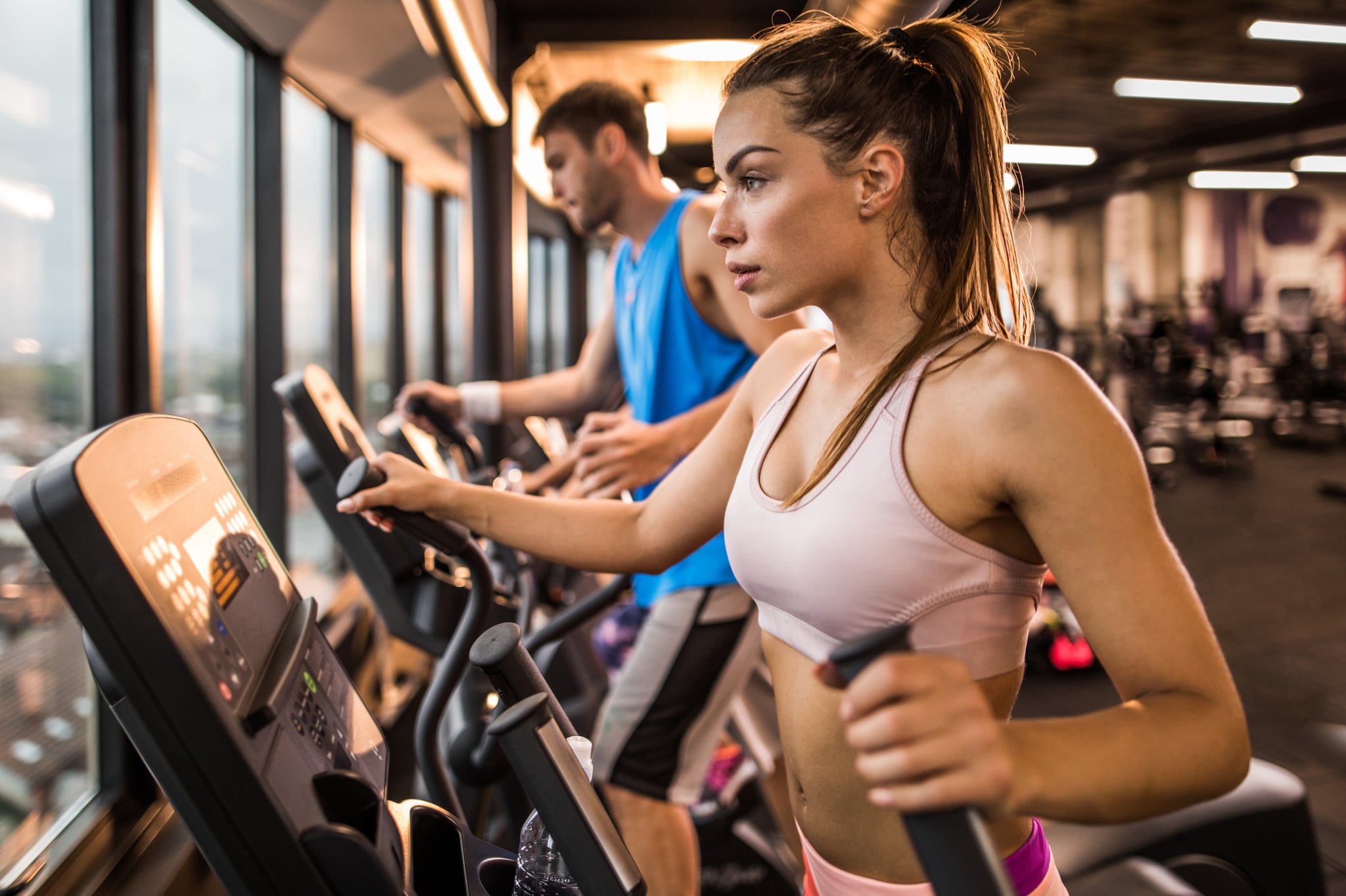 Young female athlete having a sports training on a cross trainer in a health club, while man is in the background.
