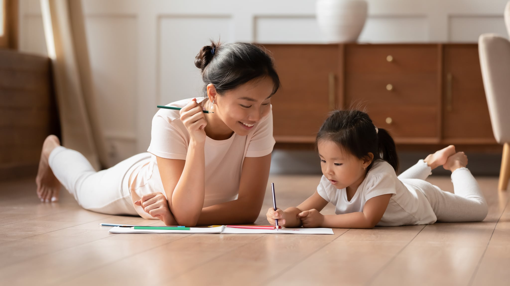 Happy young Asian mother and cute little biracial daughter lying on warm wooden floor in living room painting together, millennial ethnic mom or nanny relax with small Vietnamese girl child drawing