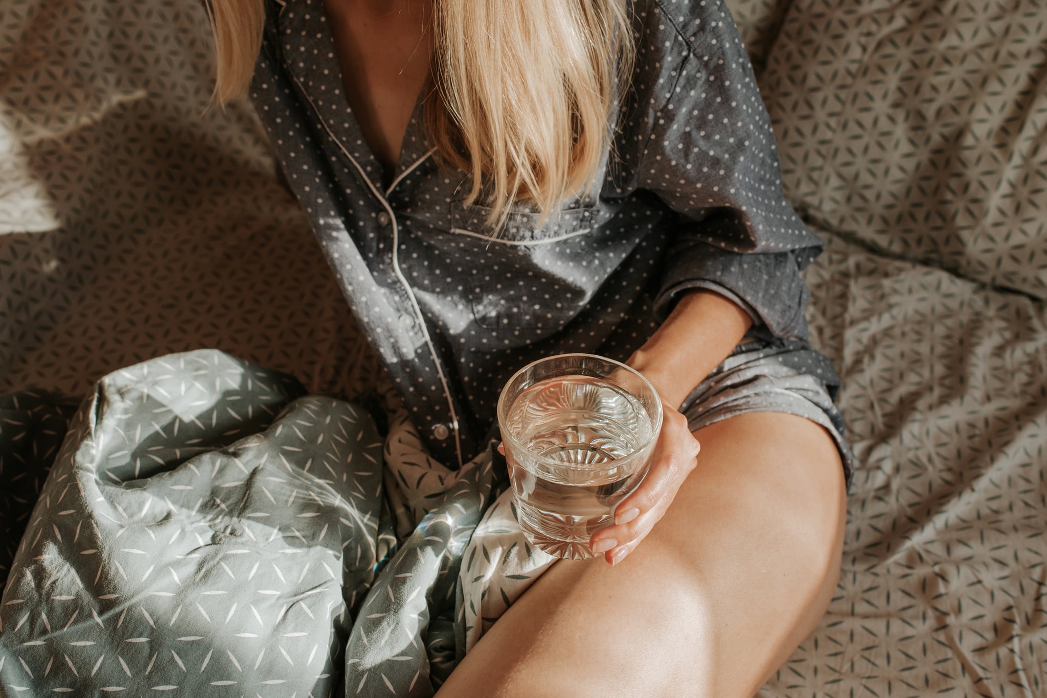 Woman in bed holding a glass of water in hand. Happy morning. Girl in pajamas. Healthy lifestyle, wellness. Proper nutrition. Drinking water. Morning with water. Sunlight on linens. Pillow, blanket