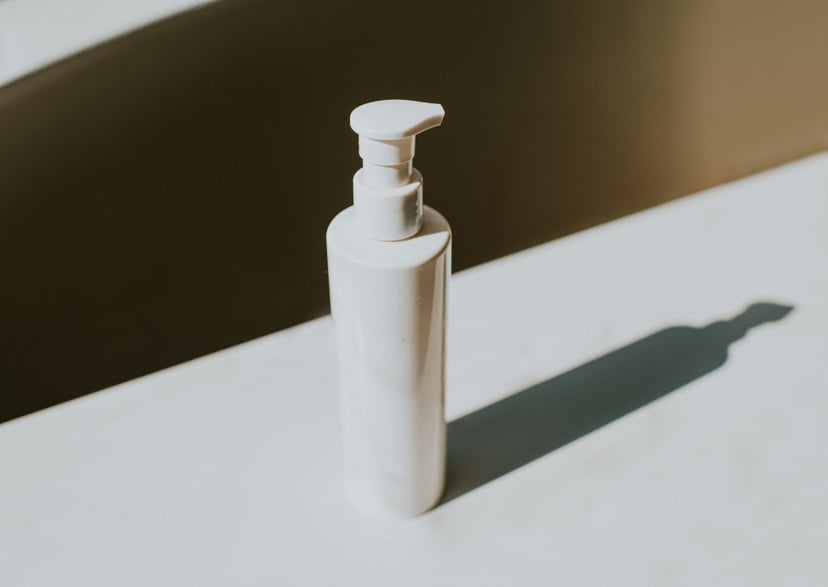 Simple conceptual image of a white plastic bottle with a pump dispenser sitting on a white surface, casting a long shadow. Space for copy.
