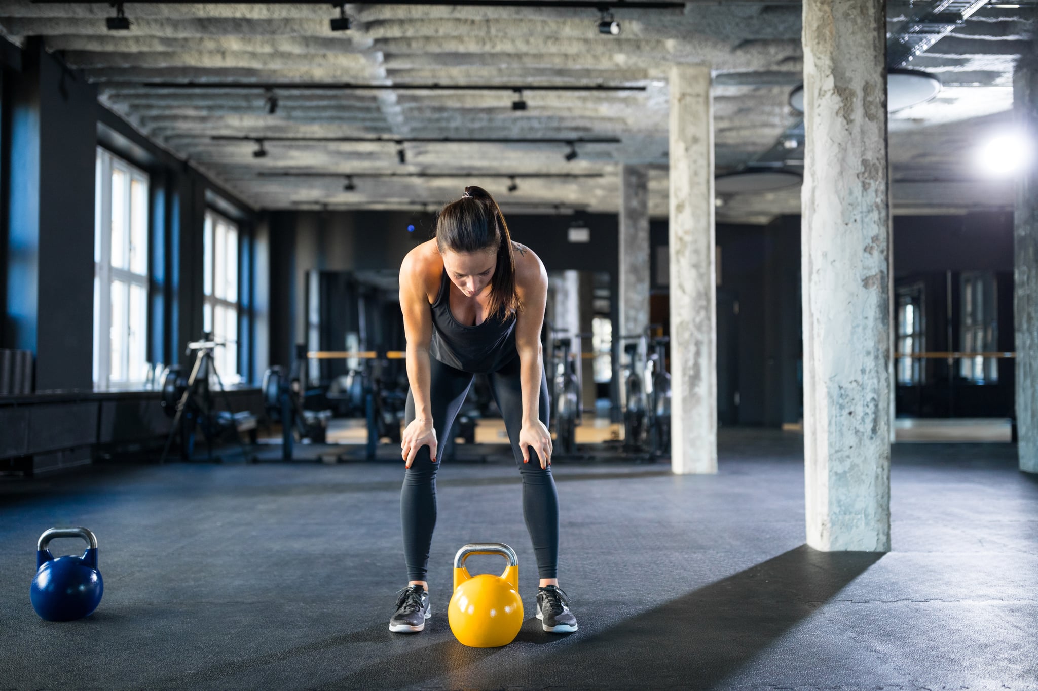 Tired female athlete standing with hands on knees in gym. Determined young woman taking a break after workout at health club. She is wearing sports clothing.