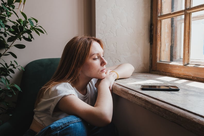 Upset redhead teen girl sitting by window looking at phone waiting call from boyfriend, feeling sad and depressed teenager looking at smartphone wait for message. Social Media depression in teens