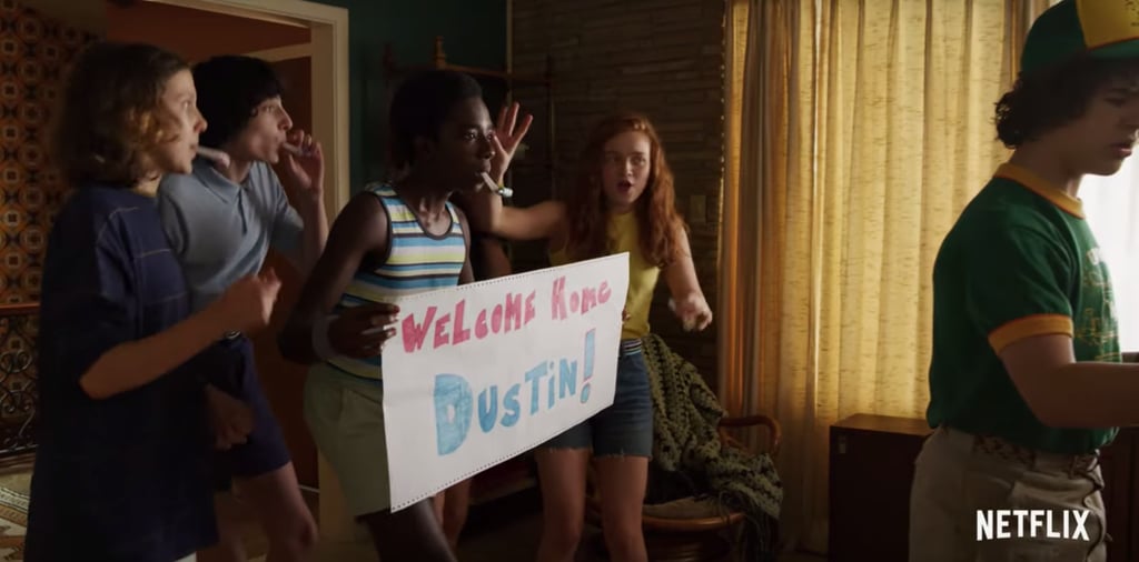 The trailer begins with the gang using Eleven's powers to help surprise Dustin, who has apparently been away for long enough that they'd go out of their way to make a "Welcome Home" banner. We assumed the season was starting at the very beginning of Summer, meaning school had only just let out, but that's clearly not the case. 
Dustin is wearing a shirt and hat that say "Camp Know Where," which has a logo covered in telescopes and computers. Apparently, he's returning home from science camp, which must mean the season is picking up halfway through what looks like a fairly normal Summer.