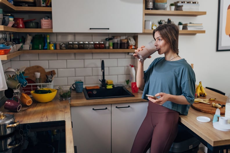 Fit woman drinking healthy fruit milkshake for breakfast while holding mobile phone in hand