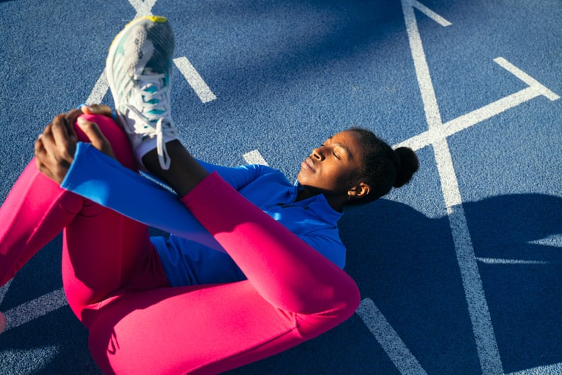 A black woman is on a blue running track stretching, wondering if she should work out with sore muscles