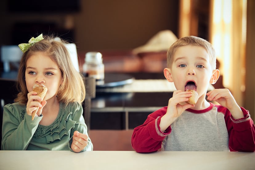 Boy and girl twins at home seated at counter in their kitchen eating ice cream cones, boy is caught wide eyed mid lick.