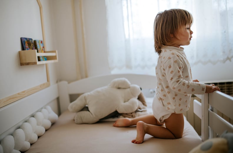 A toddler stands awake in her crib during daylight hours.