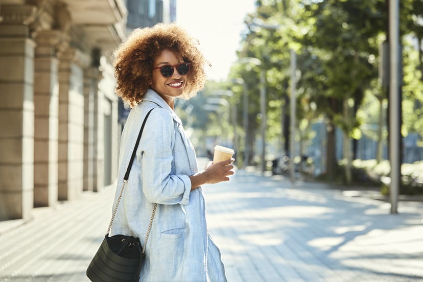Portrait of smiling young woman holding disposable coffee cup. Beautiful female is with purse walking on sidewalk. She is wearing casuals in city.