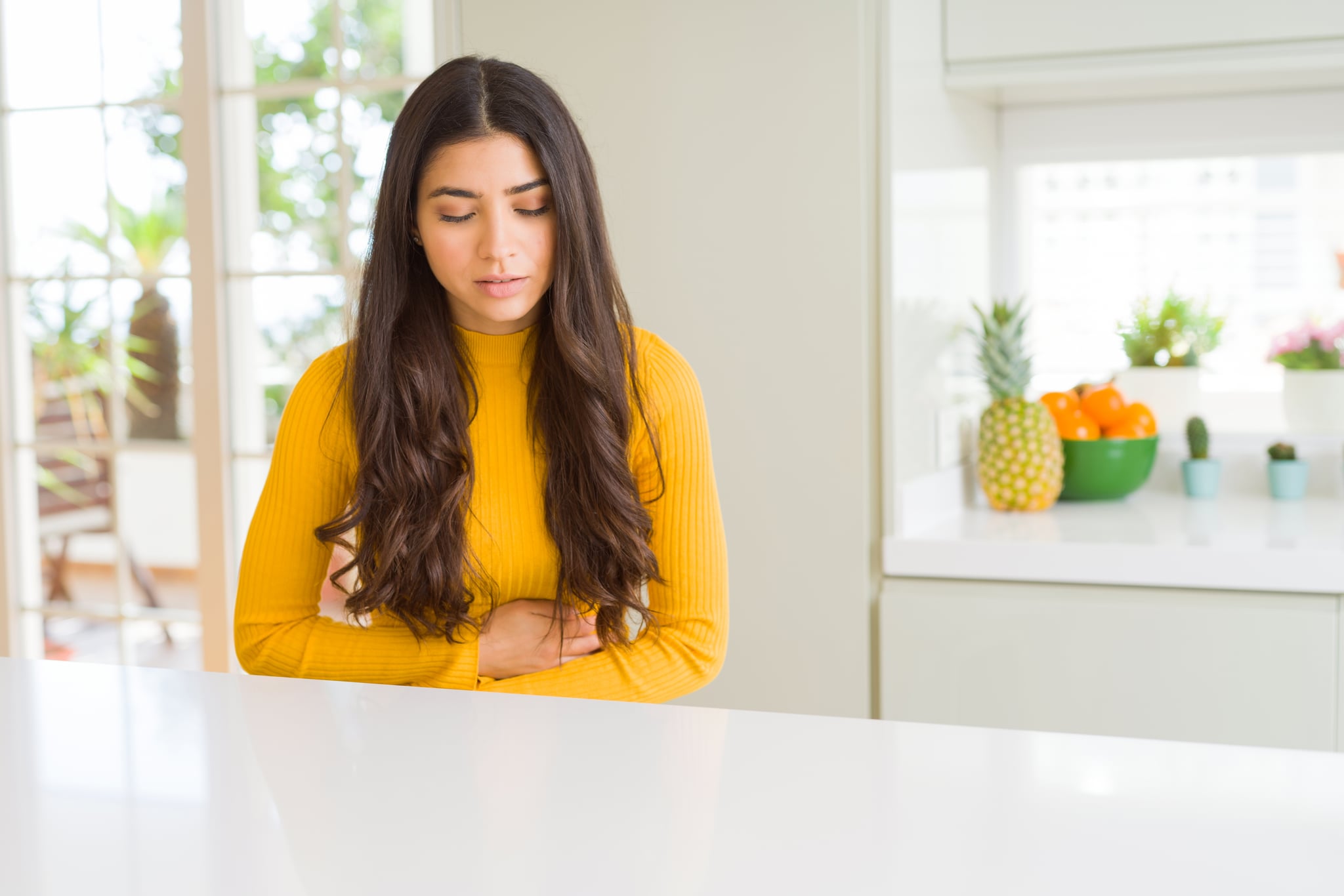 Young beautiful woman at home on white table with hand on stomach because nausea, painful disease feeling unwell. Ache concept.