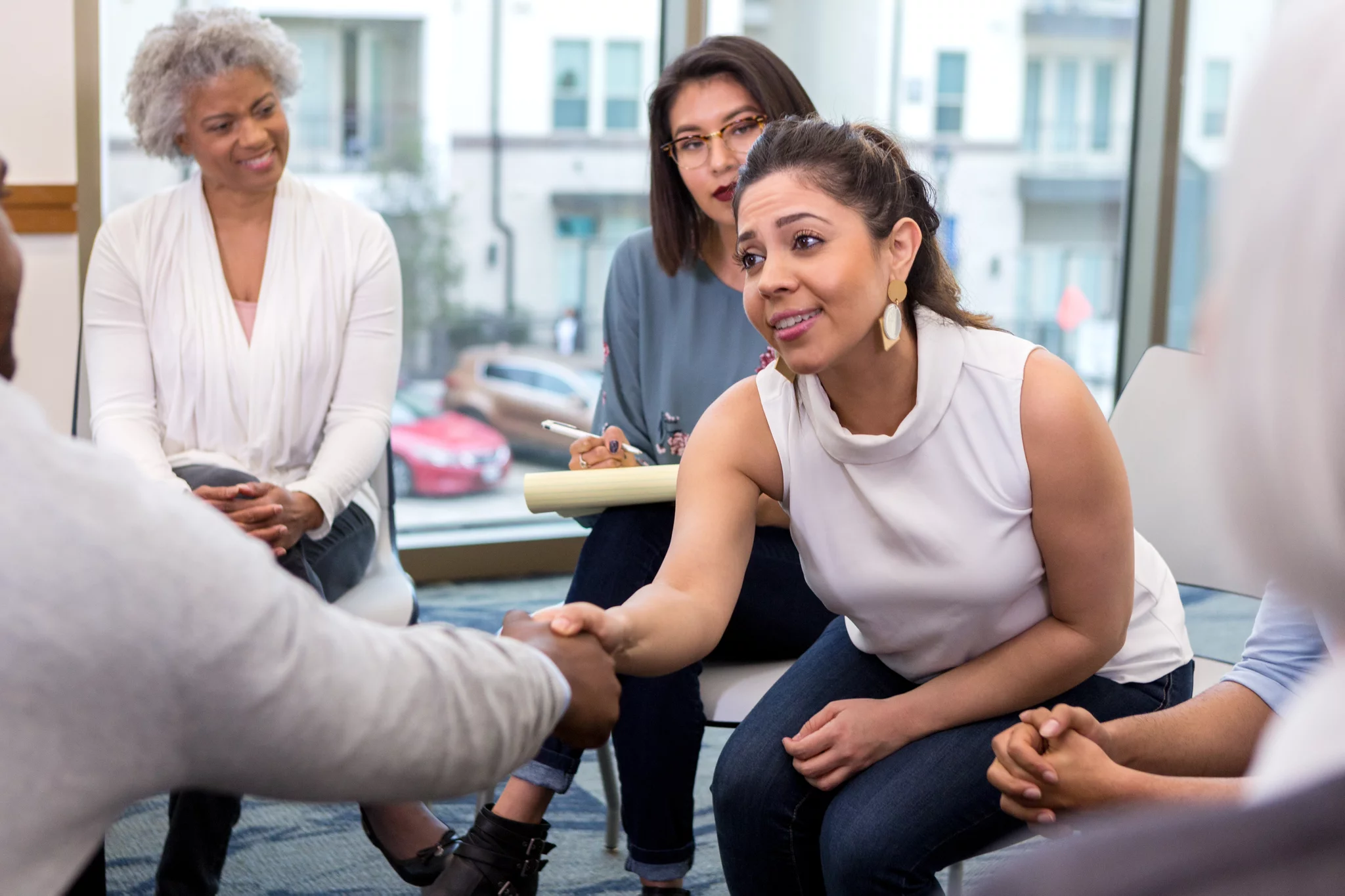 A serious young businesswoman sits with colleagues and reaches forward to shake hands with an unseen new client.