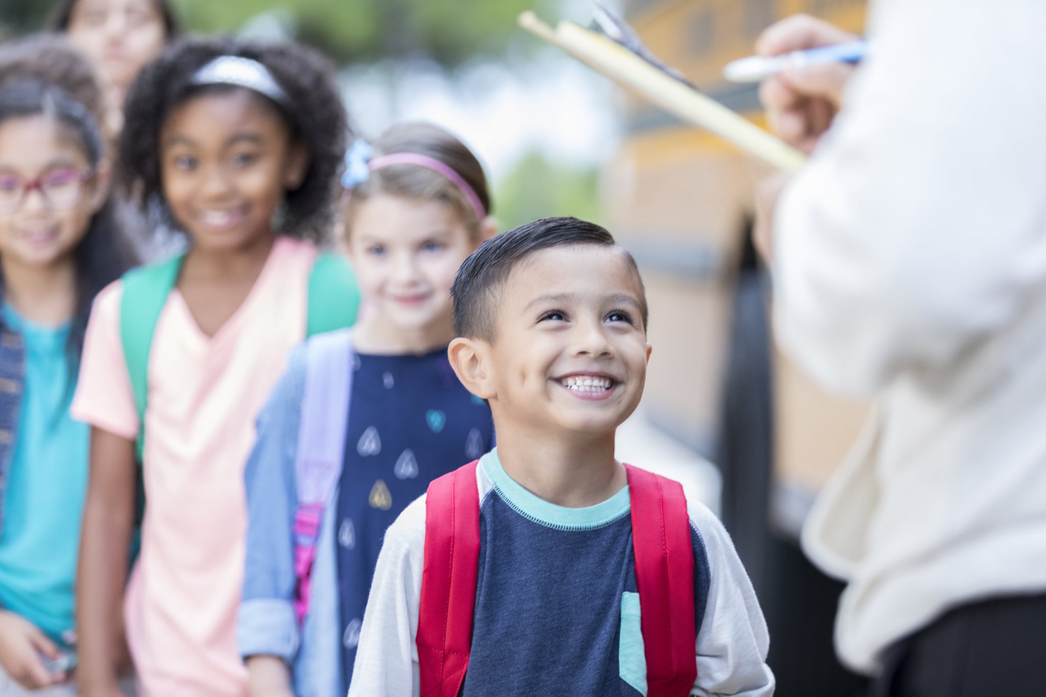 Unrecognisable school bus driver holds a clipboard and checks off each child as they get on a school bus. The children are lined up waiting to load the bus.