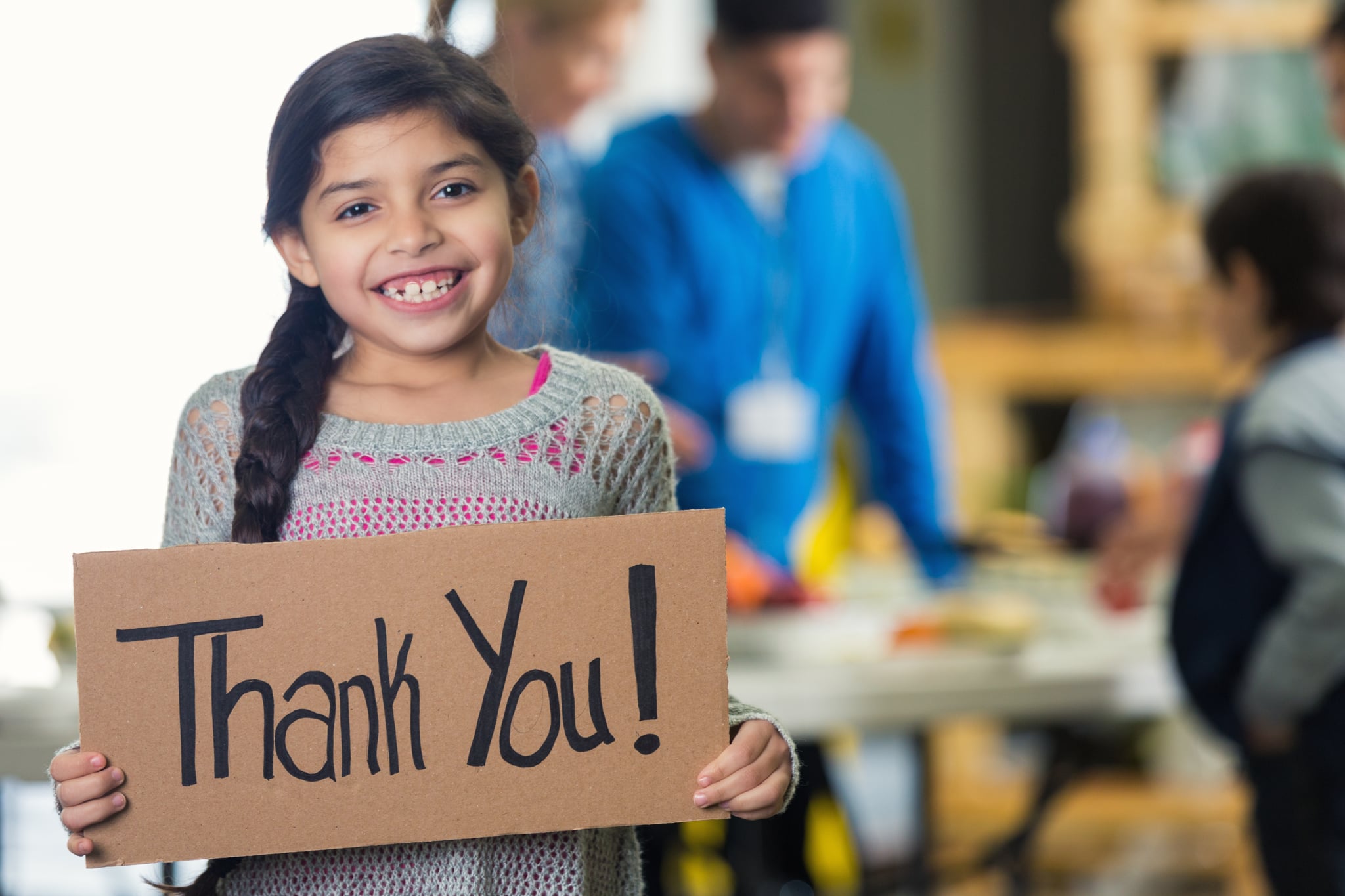 Cute Hispanic girl is holding a cardboard sign with 'Thank You!' witten on the board. She and her family are in a soup kitchen or food bank. She is smiling at the camera. Her brown hair is in a braid. Volunteers ar serving her family in the background. Focus is on the girl and the sign.