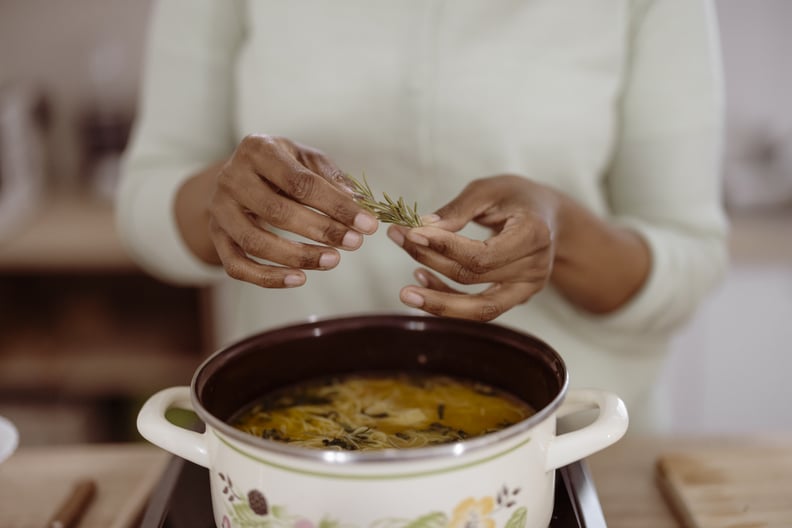 close-up of hands adding rosemary to a simmering homemade soup on the stove