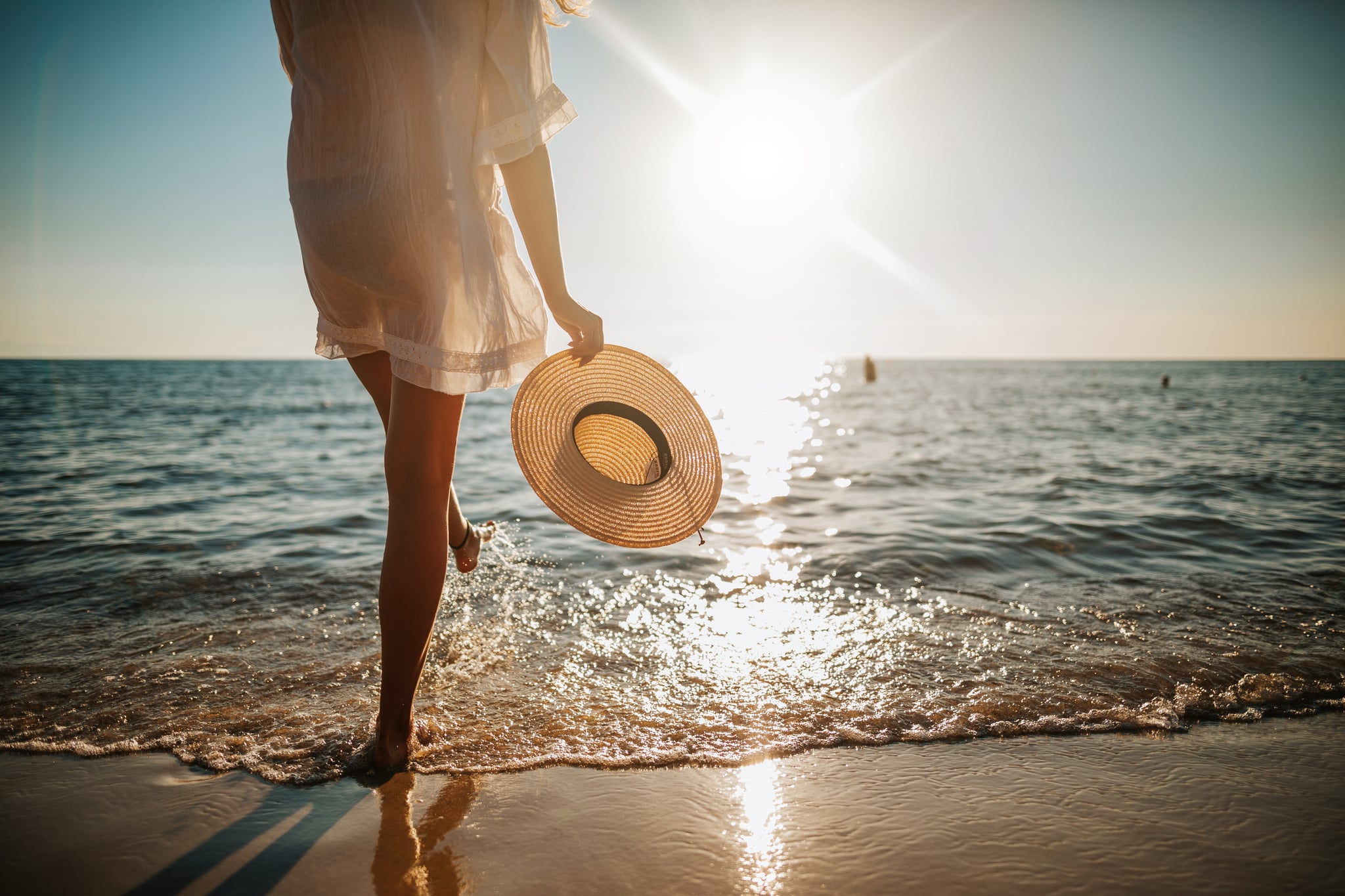 Close-up of young woman in white sun dress and with hat in hand walking alone on sandy beach at summer sunset, splashing water in sea shallow